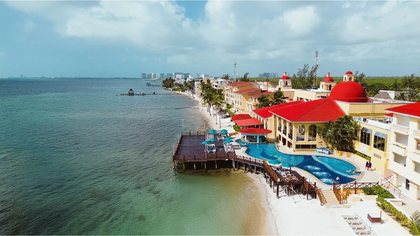 Aerial view of a beachfront resort with a swimming pool, red-roofed buildings, and palm trees along the shoreline, overlooking the ocean.