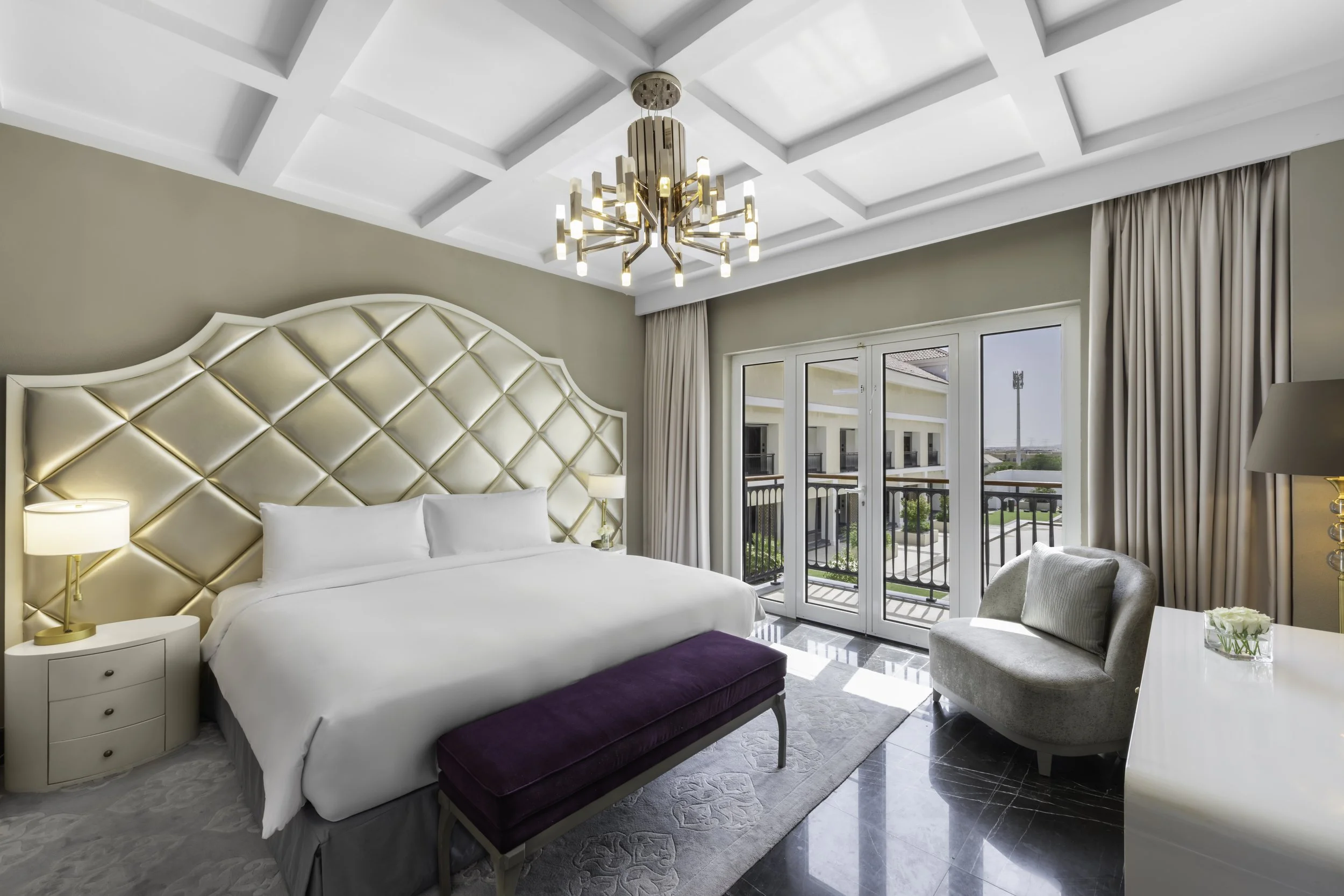 A modern bedroom with a white bed, gold and white nightstands, a tufted gold headboard, a purple bench at the foot of the bed, a white pouf chair, large windows with beige curtains, and a geometric chandelier ceiling light.
