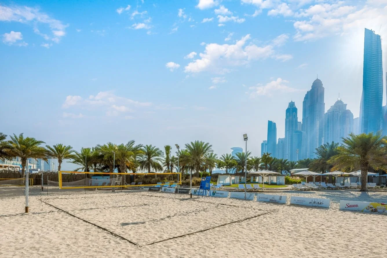 A beach volleyball court with a net, surrounded by palm trees, beachfront lounge chairs, and cabanas, with a city skyline featuring tall skyscrapers in the background on a sunny day.