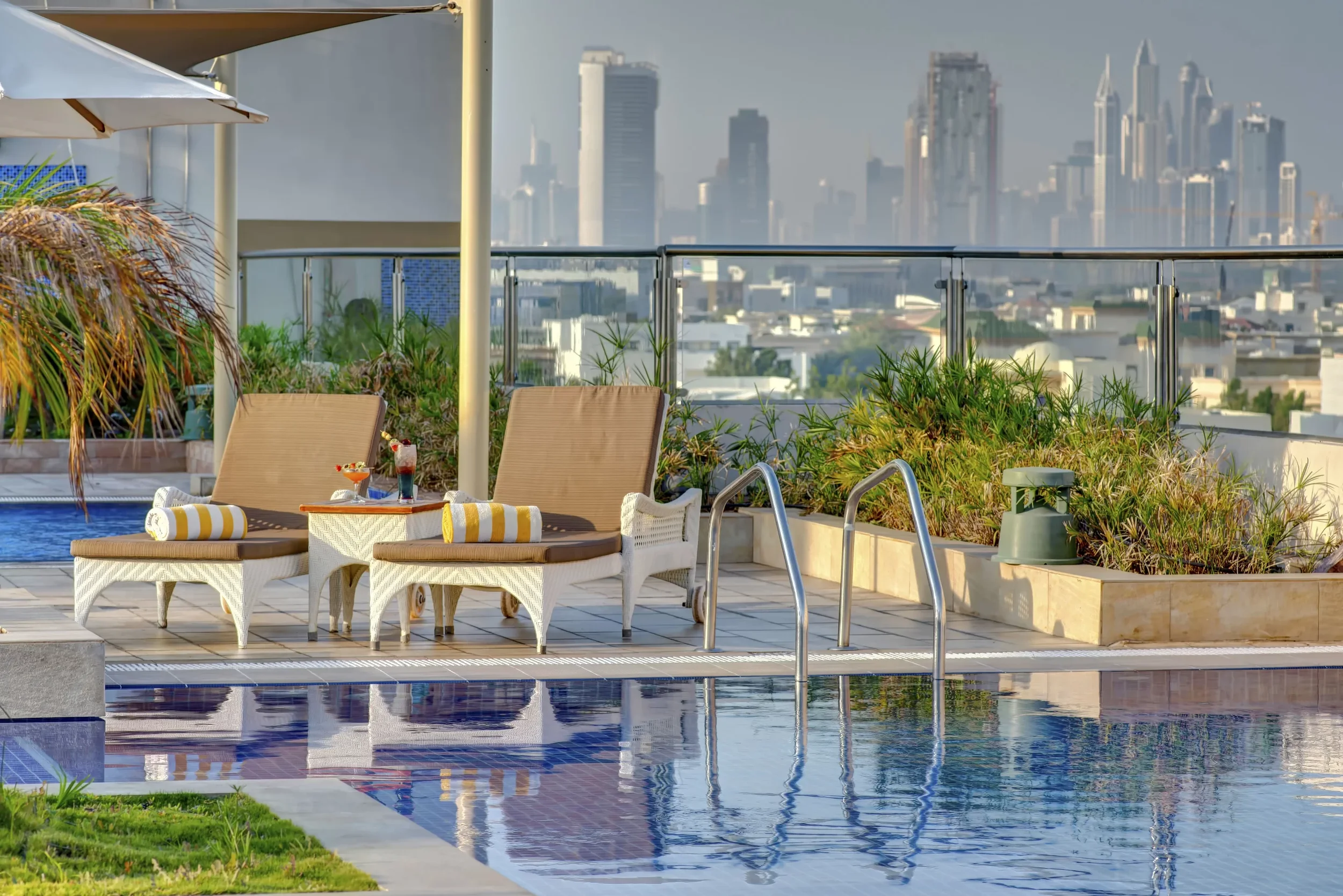 Rooftop pool area with lounge chairs, towels, drinks, and potted plants. City skyline with tall buildings in the background.