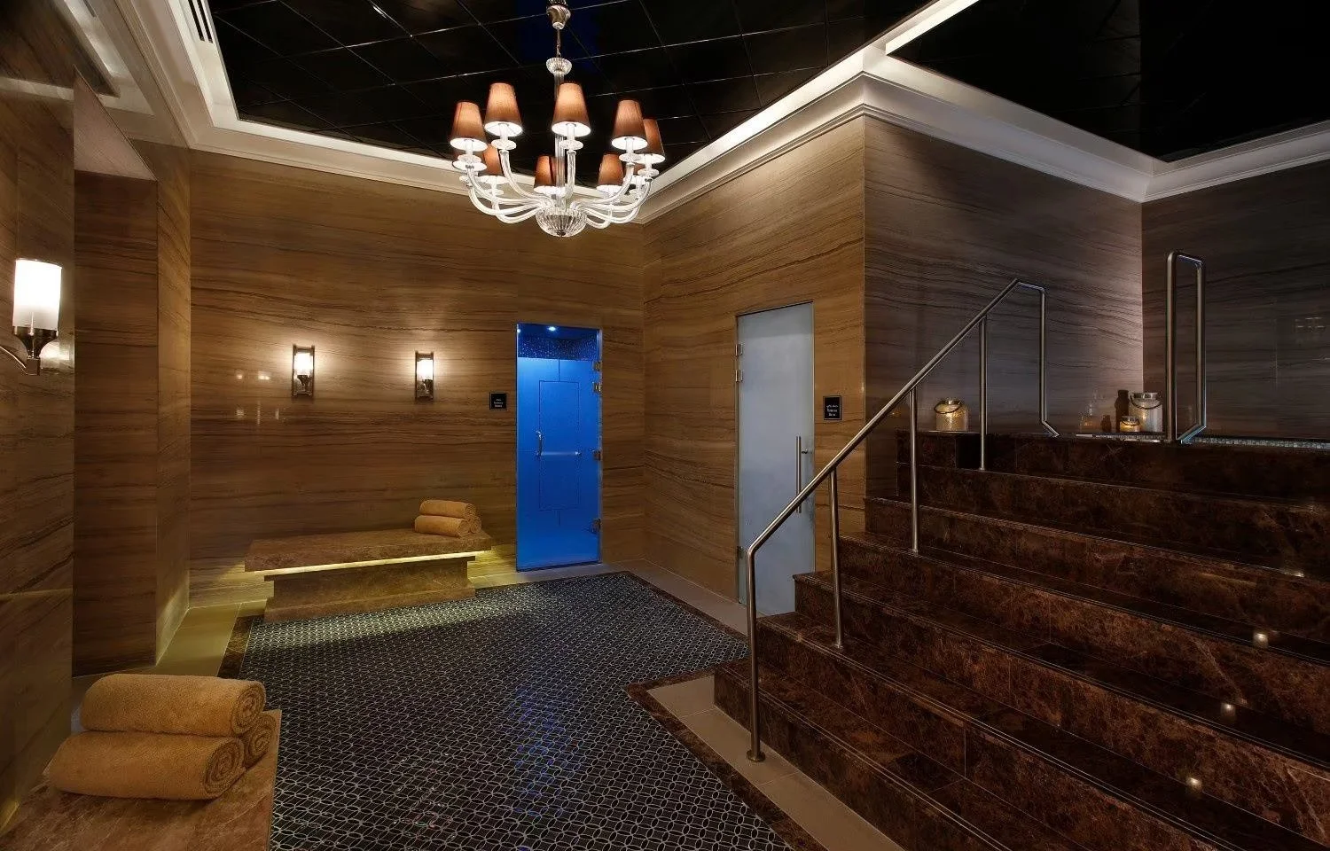 Spa area with wooden wall paneling, a chandelier, and tiled stairs leading to a hot tub with glass barriers. There are rolled towels on a bench and a floor with patterned tiles.