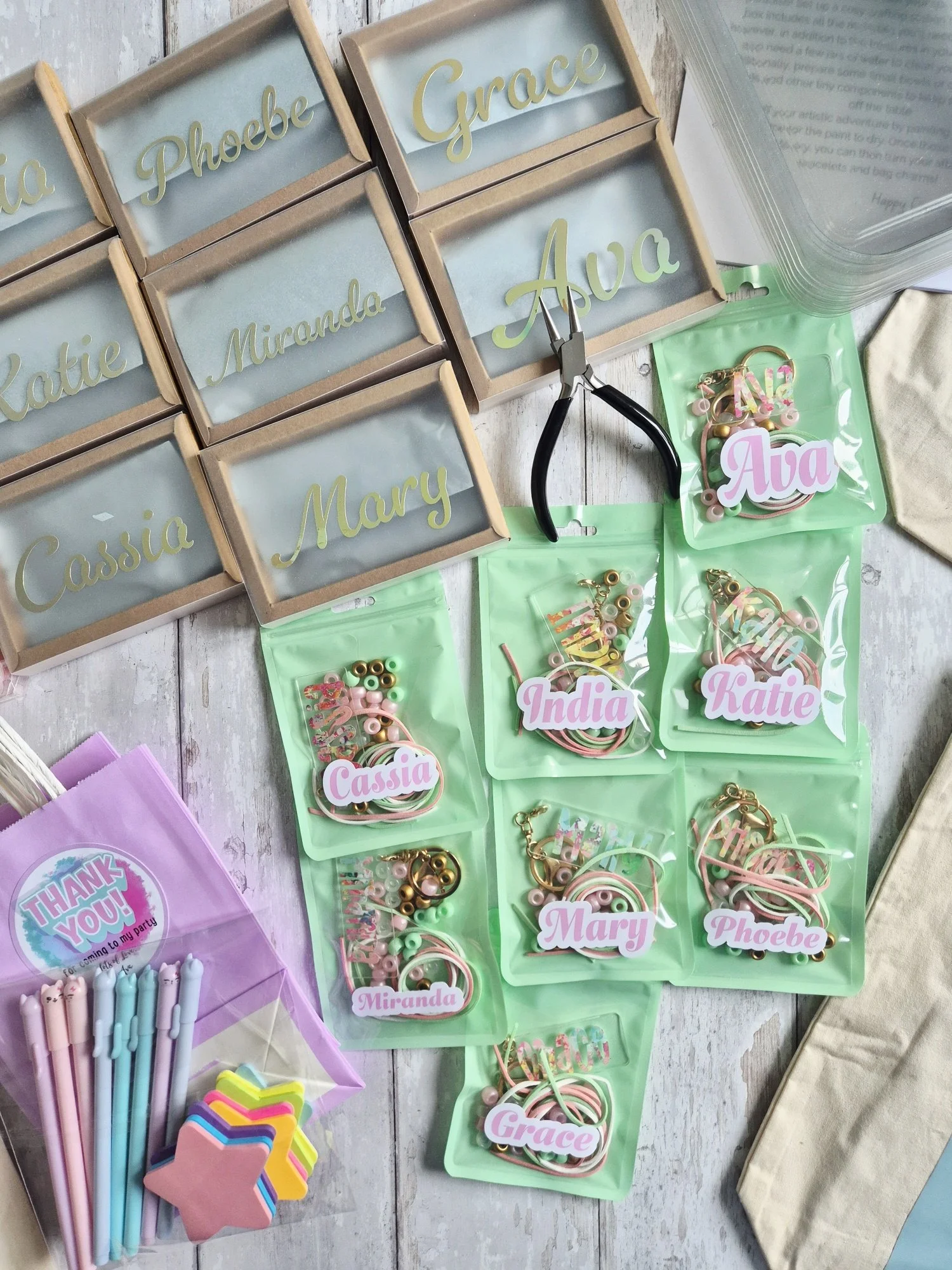 Decorative boxes with gold letter names on a wooden surface, containing small accessories and keys, and party supplies including colorful pens, straws, and star-shaped foams.
