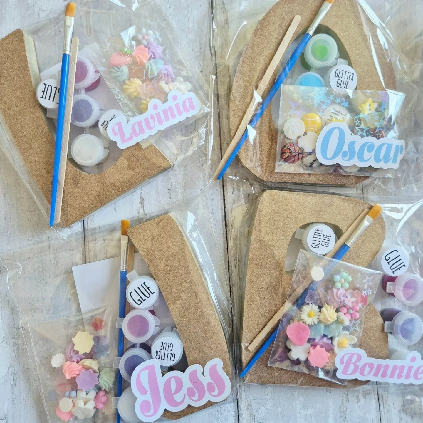 Transparent packages containing mdf letter shapes, small containers of glitter glue, paint pots, decorative foam shapes, paintbrushes, and name labels for girls named Lavinia, Oscar, Jess, and Bonnie.