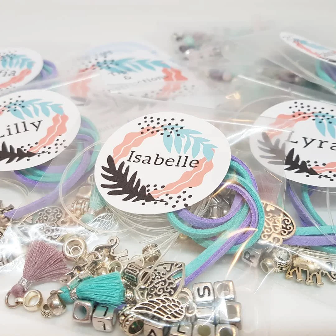 Multiple small bags of personalised bracelets making materials and charms labelled with the name 'Isabelle' with straw-coloured elastic bands, tassels, and metallic charms including hearts, the word 'Love,' and other decorative shapes.