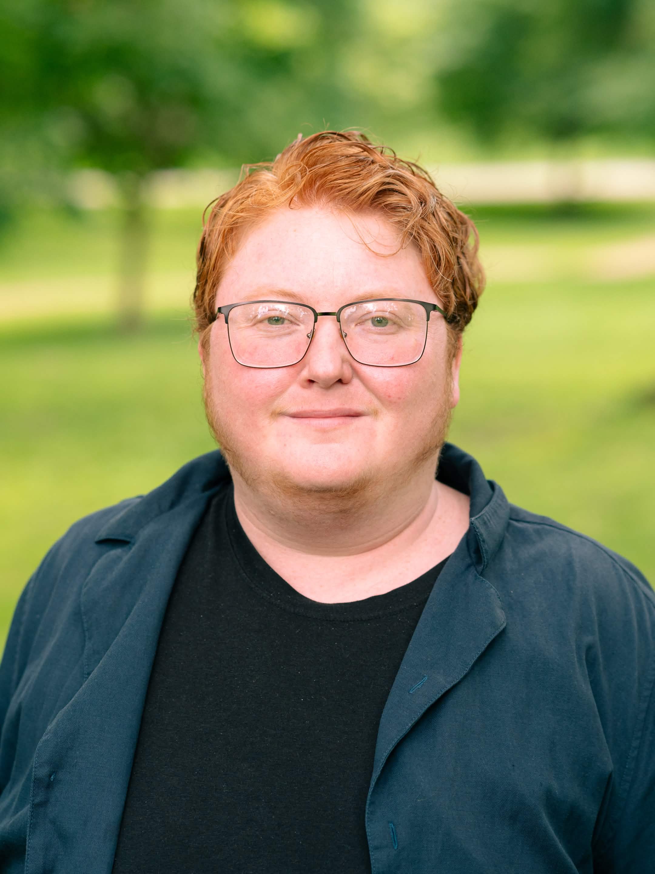 A person with red hair and glasses outdoors, wearing a dark shirt and a blue jacket, with a blurred green background.