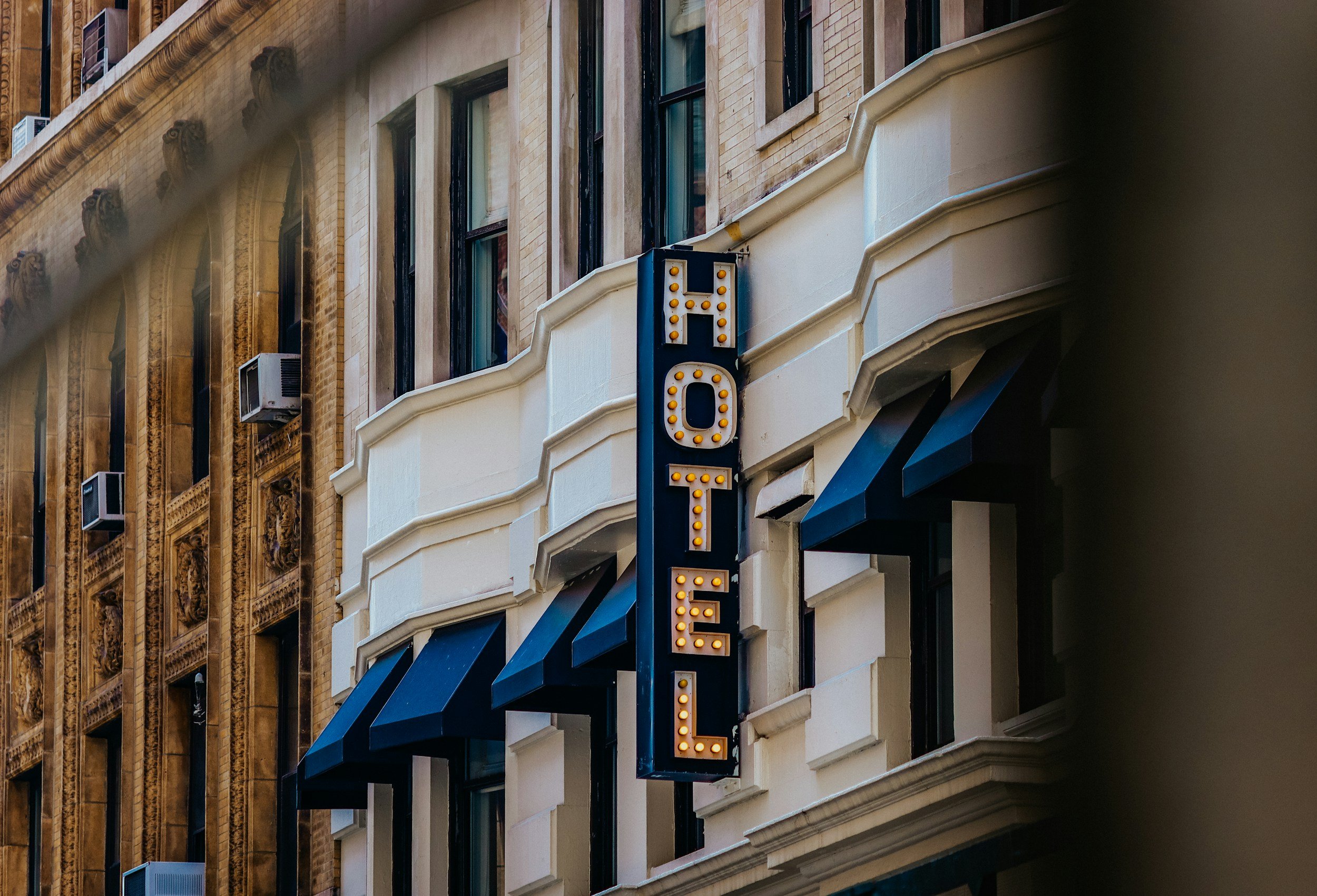 A building facade with multiple rows of windows and decorative architectural details, featuring a vertical neon sign with the word 'HOTEL' in yellow lights.