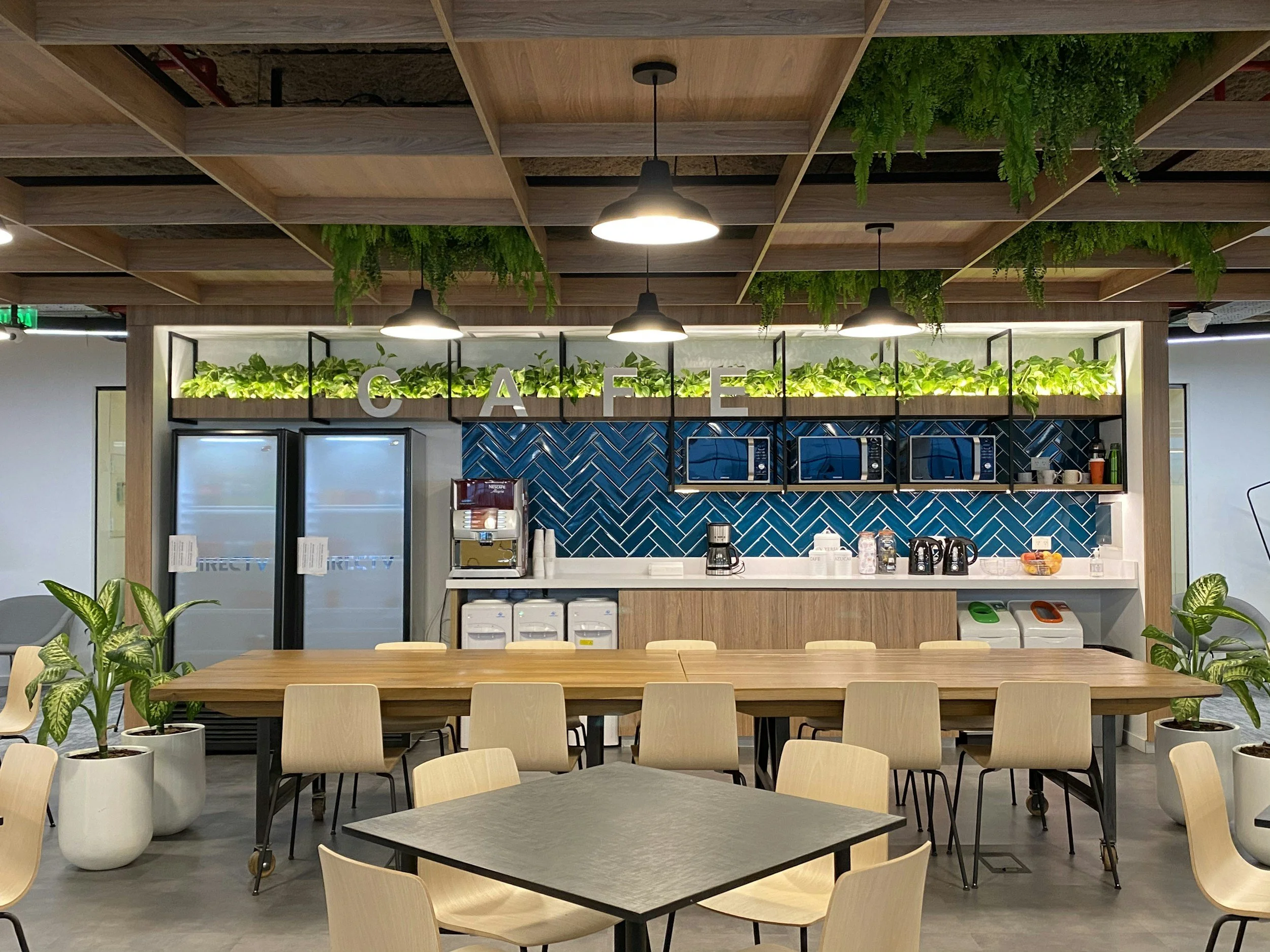 Interior of a modern cafe with a wooden ceiling, green plants, and a counter with coffee machines and microwaves, decorated with large white letters spelling 'CAFE'.