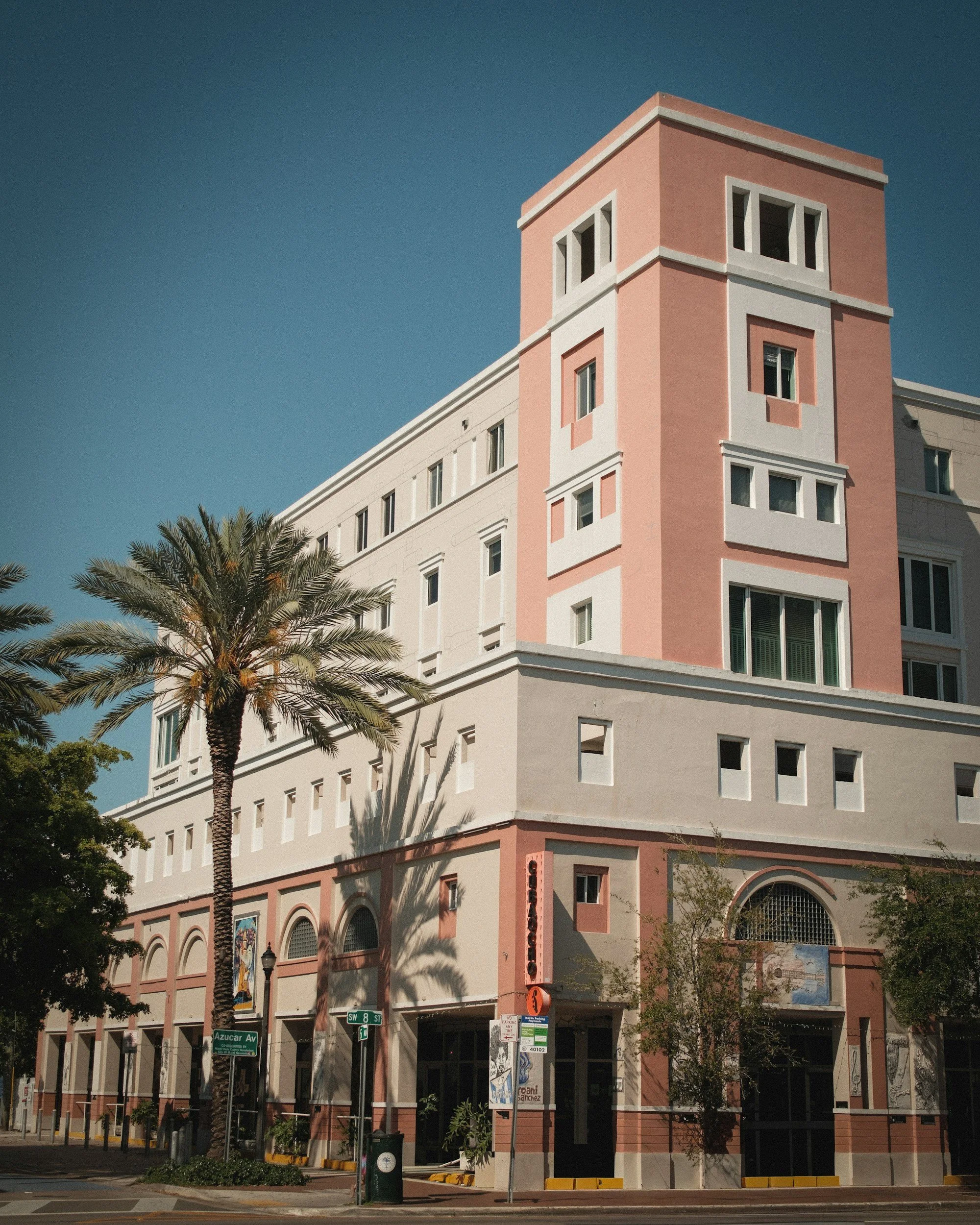 A pink and white multi-story building with rectangular windows, a palm tree in front, and street signs at the corner of SW 8th Street and Azucar Avenue.