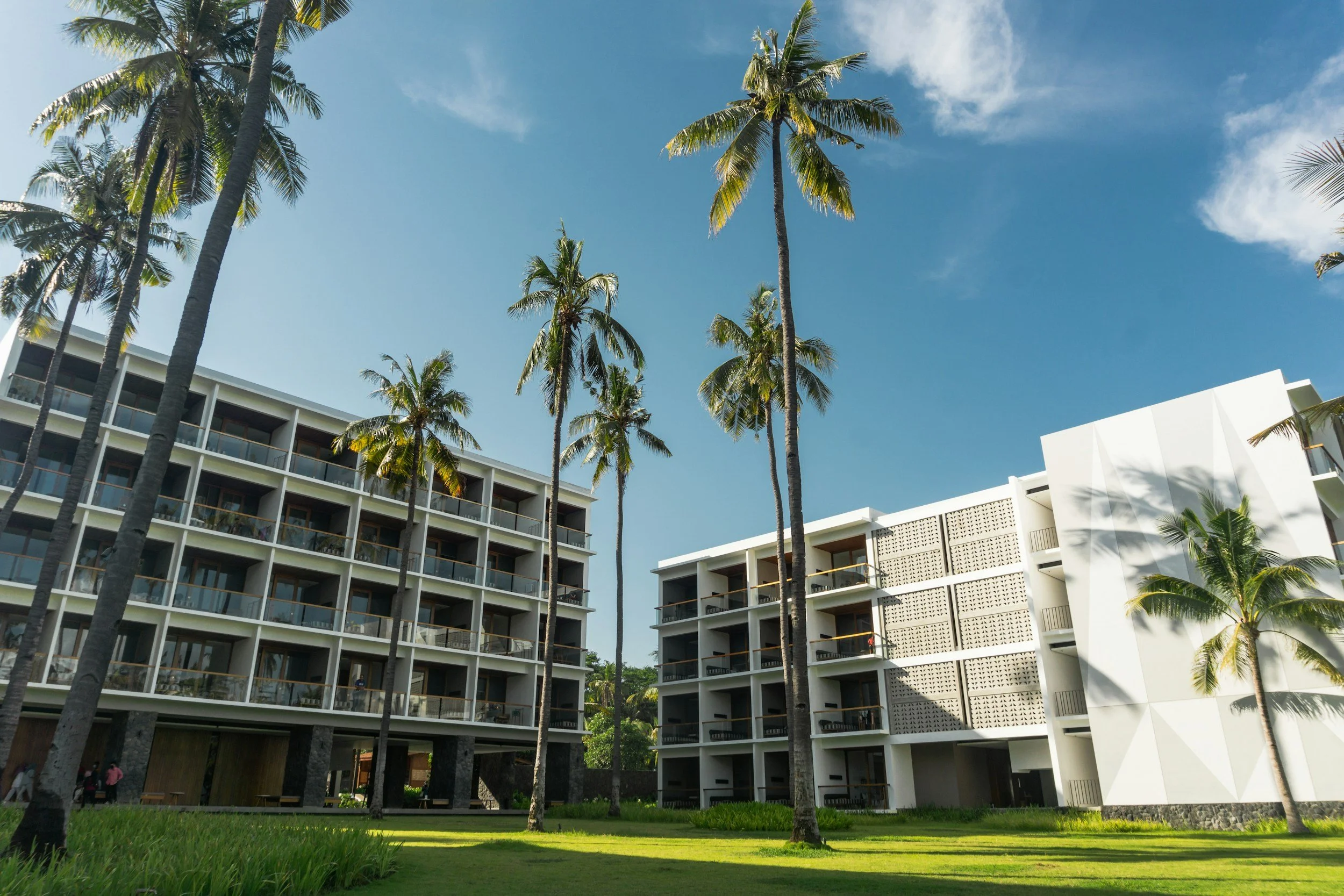 Multiple modern white buildings with balconies are surrounded by tall palm trees under a clear blue sky with some clouds.