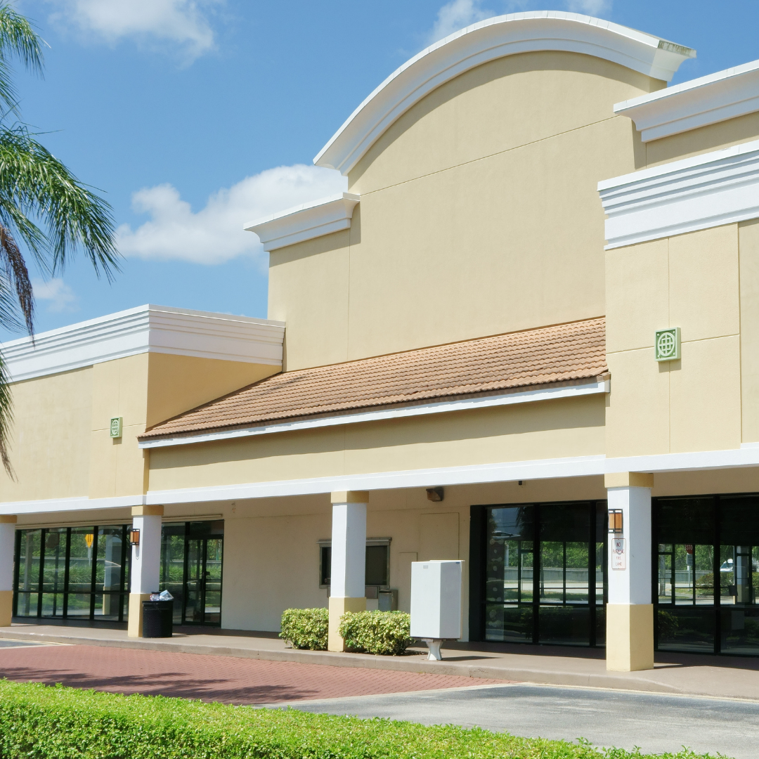 Commercial building with beige walls, large windows, and a tiled roof, under a blue sky with some clouds.