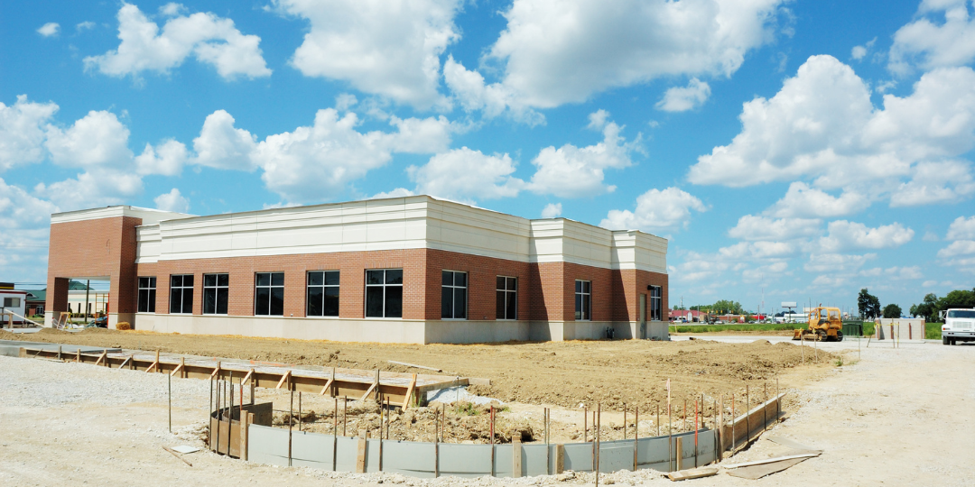 Construction site with a new building under construction, dirt ground, and construction equipment, under a partly cloudy sky.