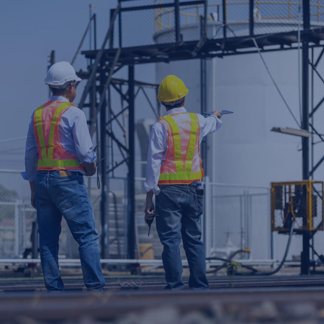Two construction workers wearing white and yellow safety helmets and high-visibility vests inspecting an industrial site with metal scaffolding and power equipment.