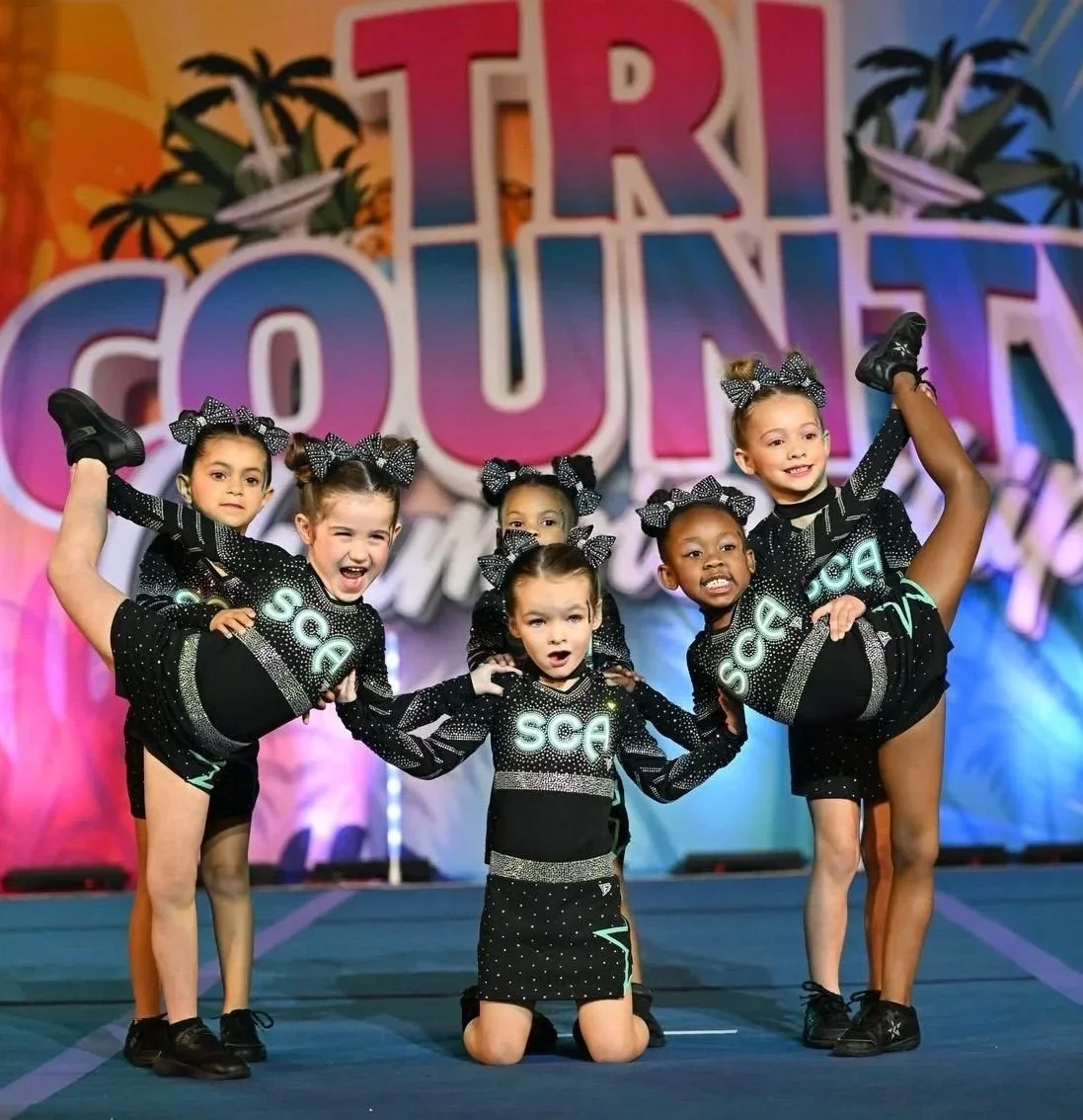 A group of six young girls in cheerleading costumes with black and neon green accents, performing a cheer routine on stage at a cheerleading competition called 'Trout Covered.' They are posing with some girls holding their legs up, showing flexibility and cheerleading skills, with a colorful backdrop featuring the event name and palm trees.