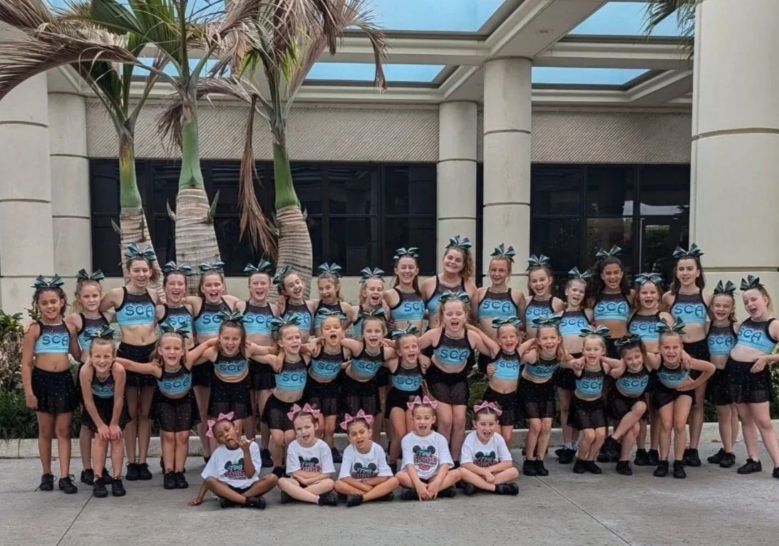 Group of young cheerleaders in matching uniforms and bows, posing in front of palm trees and a building with large windows.