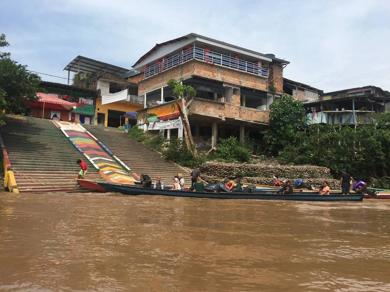 People boarding boats at the riverbank with colorful buildings and a tiled staircase leading to houses above.