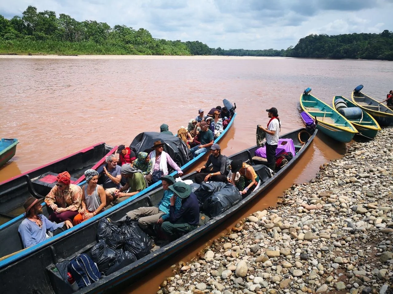 People aboard several boats on a riverbank, with green trees in the background, preparing for a boat trip, carrying bags and supplies.