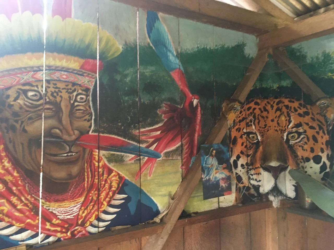 Wall mural depicting a Native American man with a feathered headdress and traditional clothing, a colorful bird with a long beak, and a jaguar's face, with a small religious picture hanging on a wooden structure.