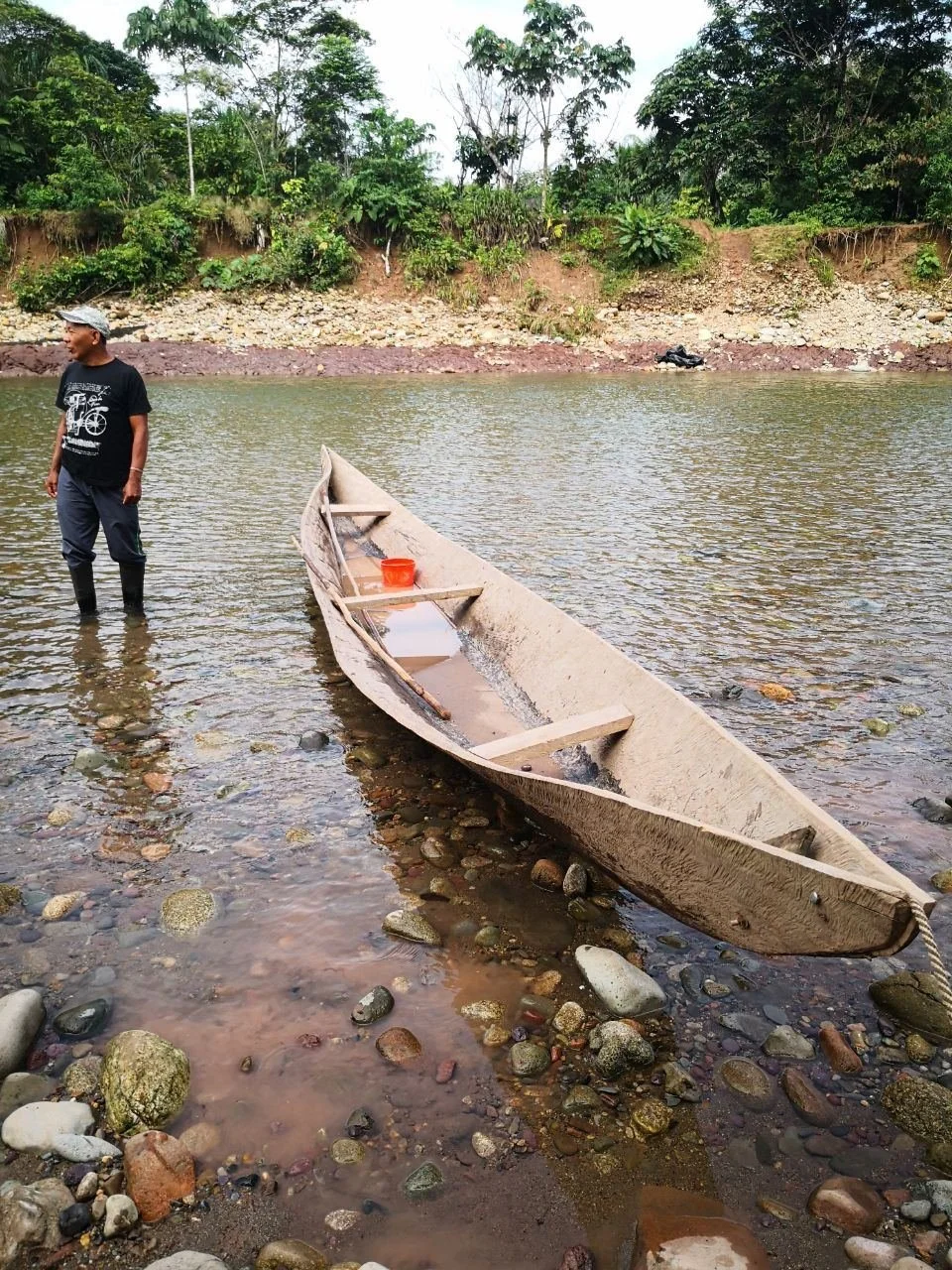 A man standing in shallow water next to a small wooden boat with a red bucket inside, on a riverbank with greenery and rocks in the background.