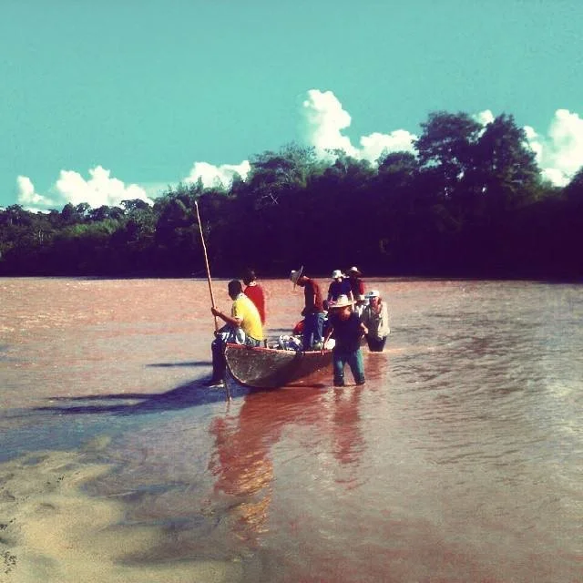 Group of people wading through a muddy river with a wooden boat, lush green trees in the background, on a sunny day with partly cloudy sky. Colombia, south America, natural medicine