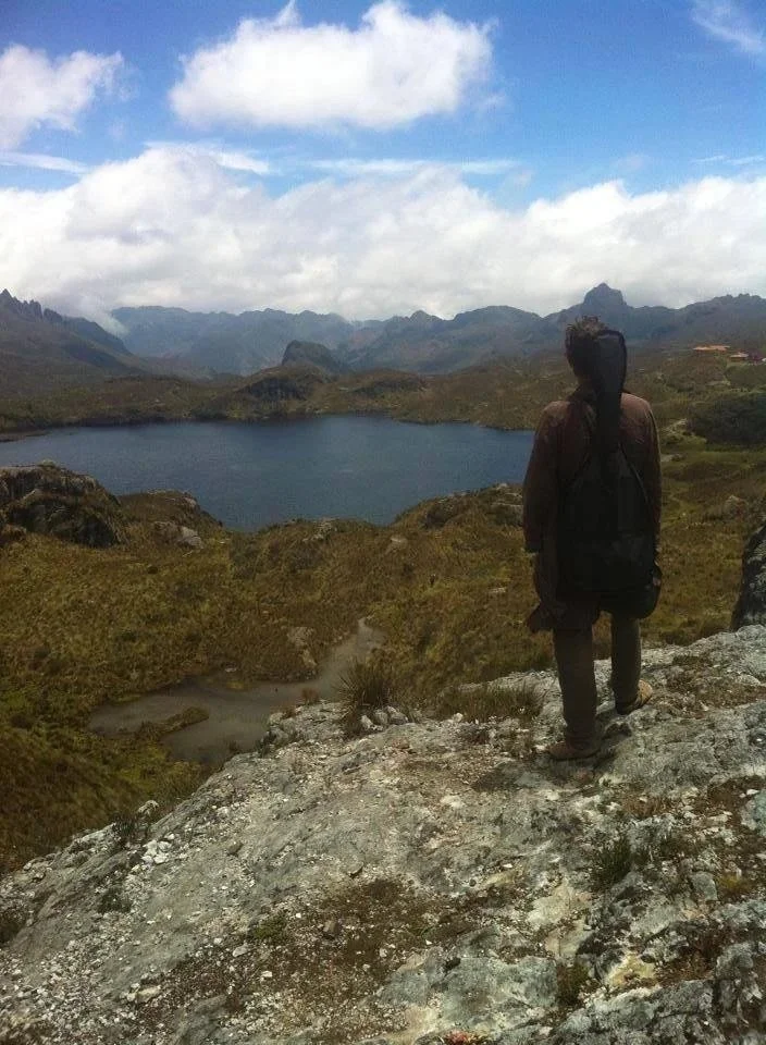 Person standing on rocky outcrop overlooking a lake and mountain range under partly cloudy sky. Chaka, Medicine, Life, Path, Freedom, Choice, Vitality,