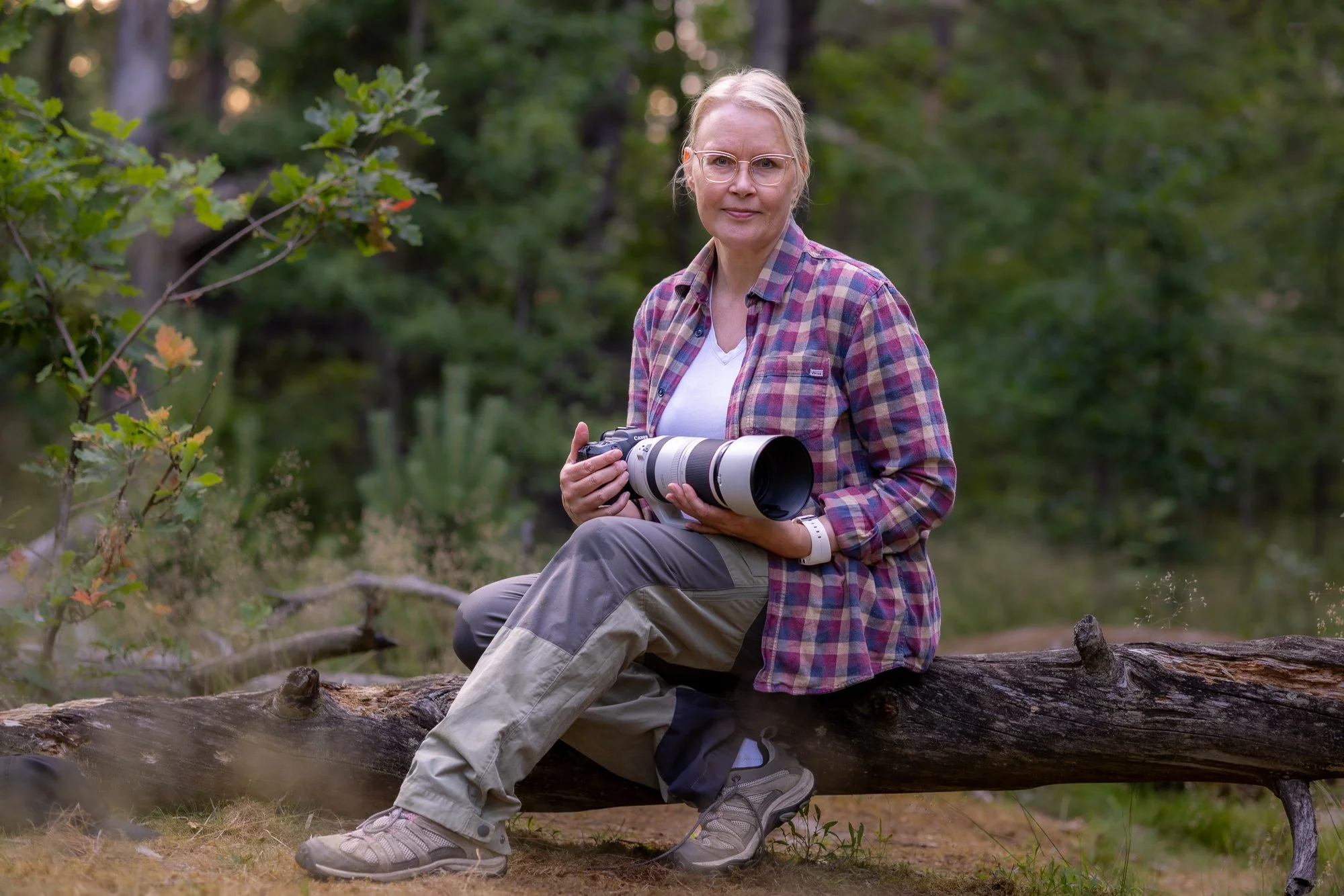 A woman with glasses, wearing a plaid shirt, sits on a log in a forest, holding a camera with a large lens.