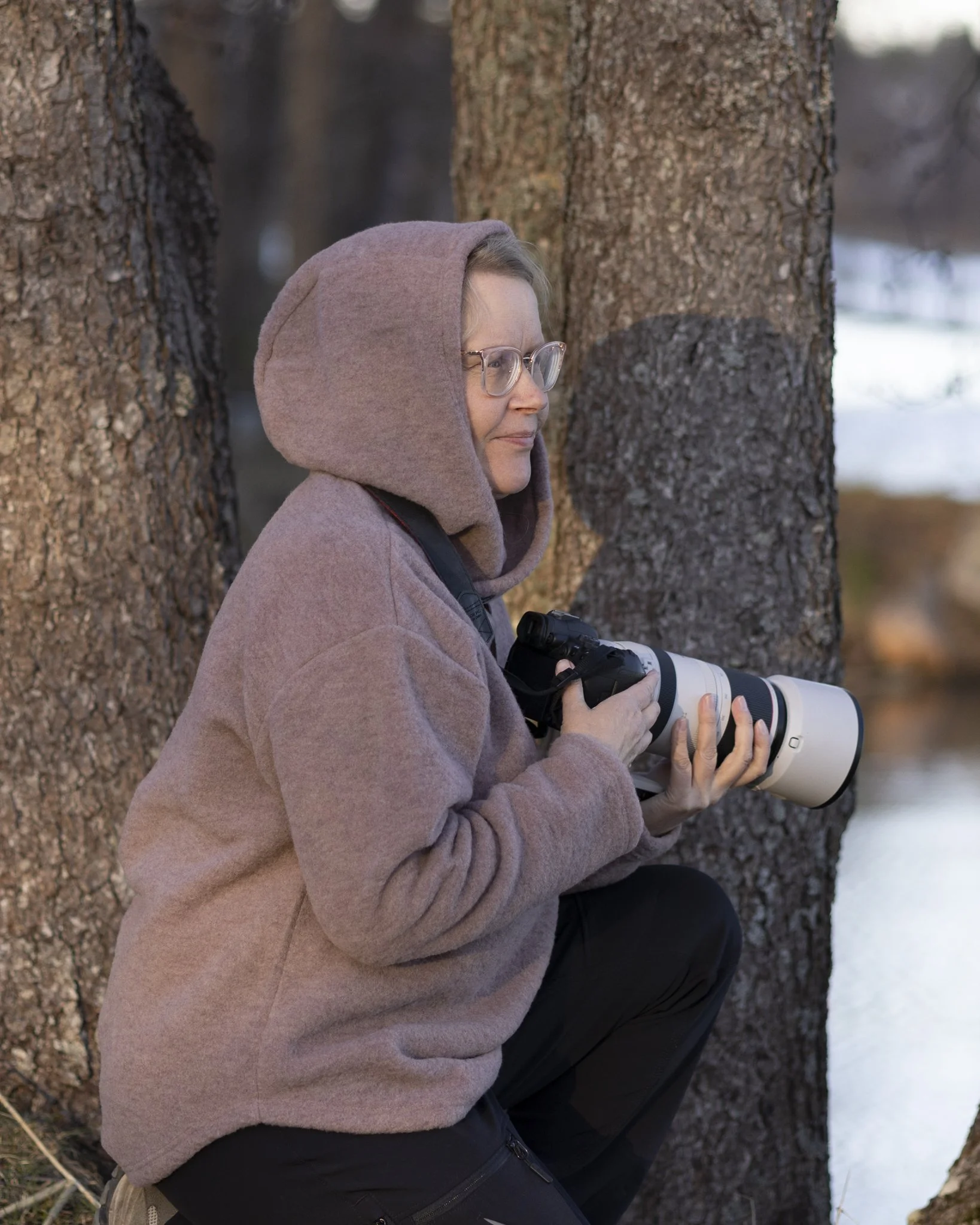 A person with glasses, wearing a brown hoodie, sitting against a tree in an outdoor setting with snow on the ground, holding a camera with a large lens.