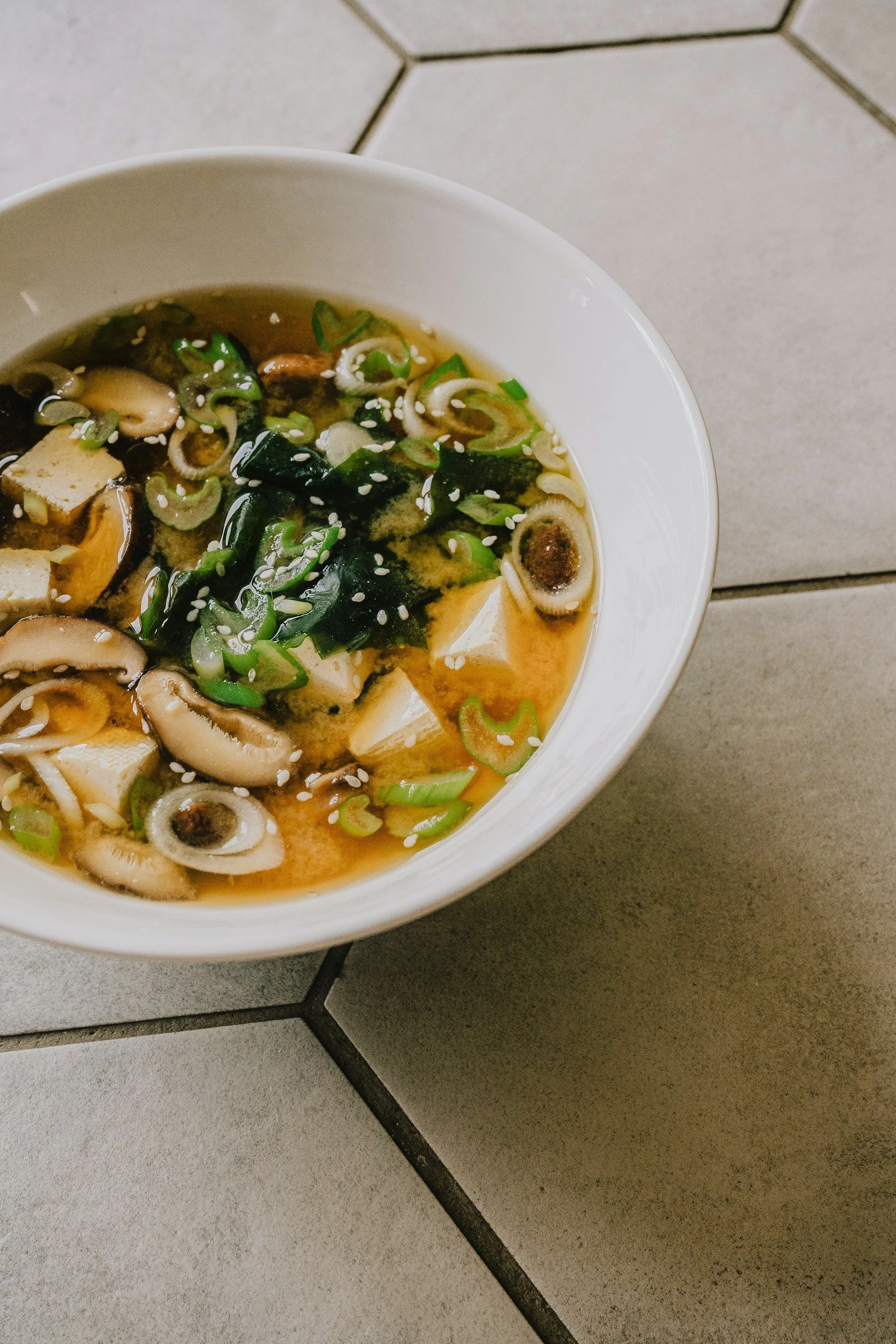 A bowl of miso soup with green onions, seaweed, tofu, mushrooms, and sesame seeds, placed on a tiled surface.