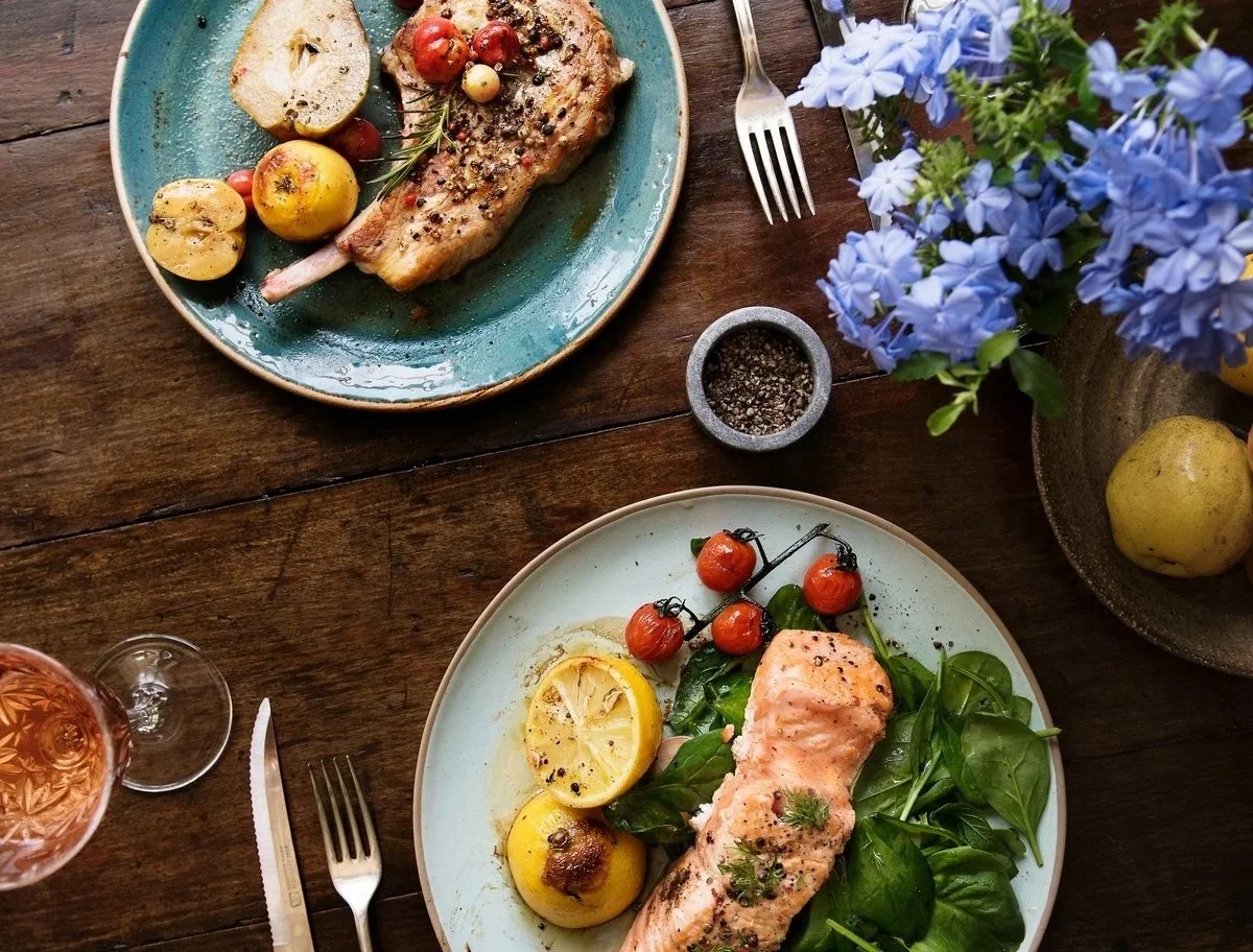A rustic wooden table with two plates of cooked salmon, lemon slices, cherry tomatoes, and green leafy vegetables, accompanied by a glass of rosé wine, a fork, a knife, a small bowl of black pepper, and a blue flower bouquet.