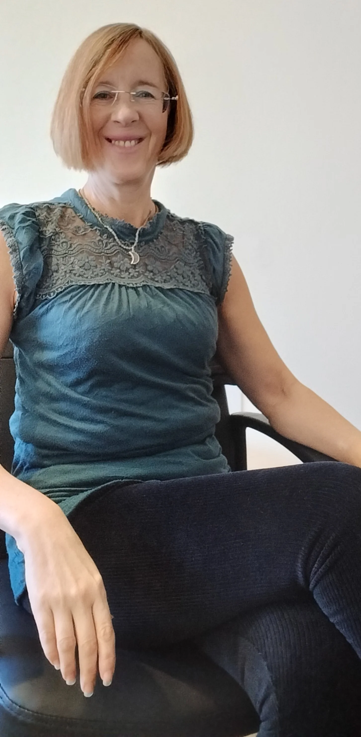 A smiling woman with short blonde hair, glasses, and a necklace sitting in a black chair against a plain white wall. She is wearing a teal blouse with lace and embroidery details and black pants.