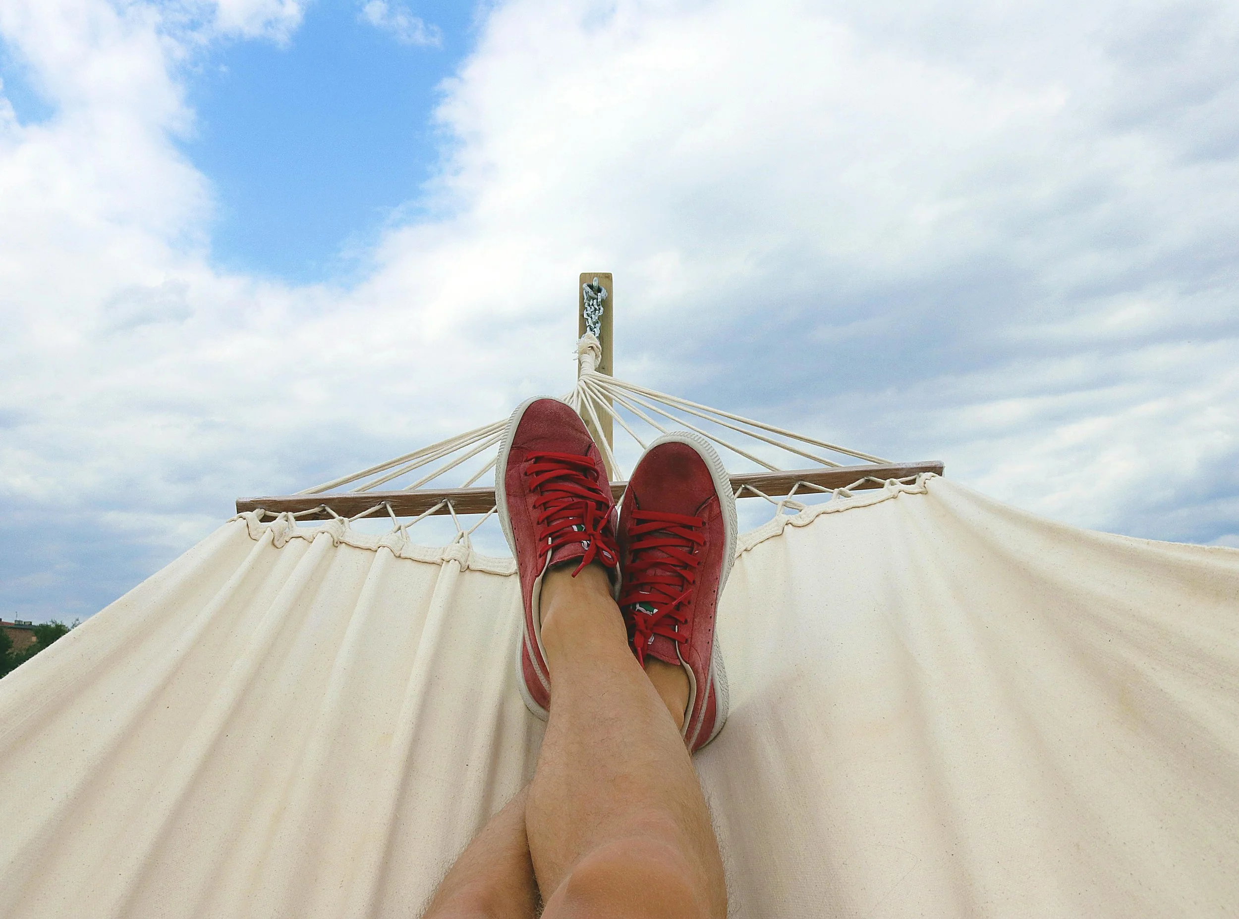 Person's legs wearing red sneakers resting on the edge of a hammock, looking up at a partly cloudy sky