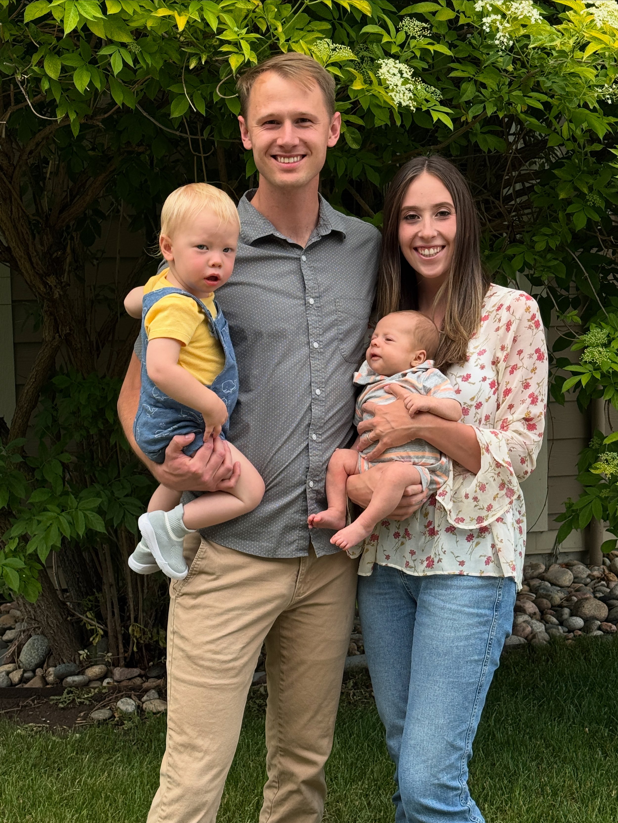 A family of four standing outdoors in front of a green leafy bush. The father is holding a young boy in a yellow shirt, and the mother is holding a baby in a striped outfit. All are smiling.