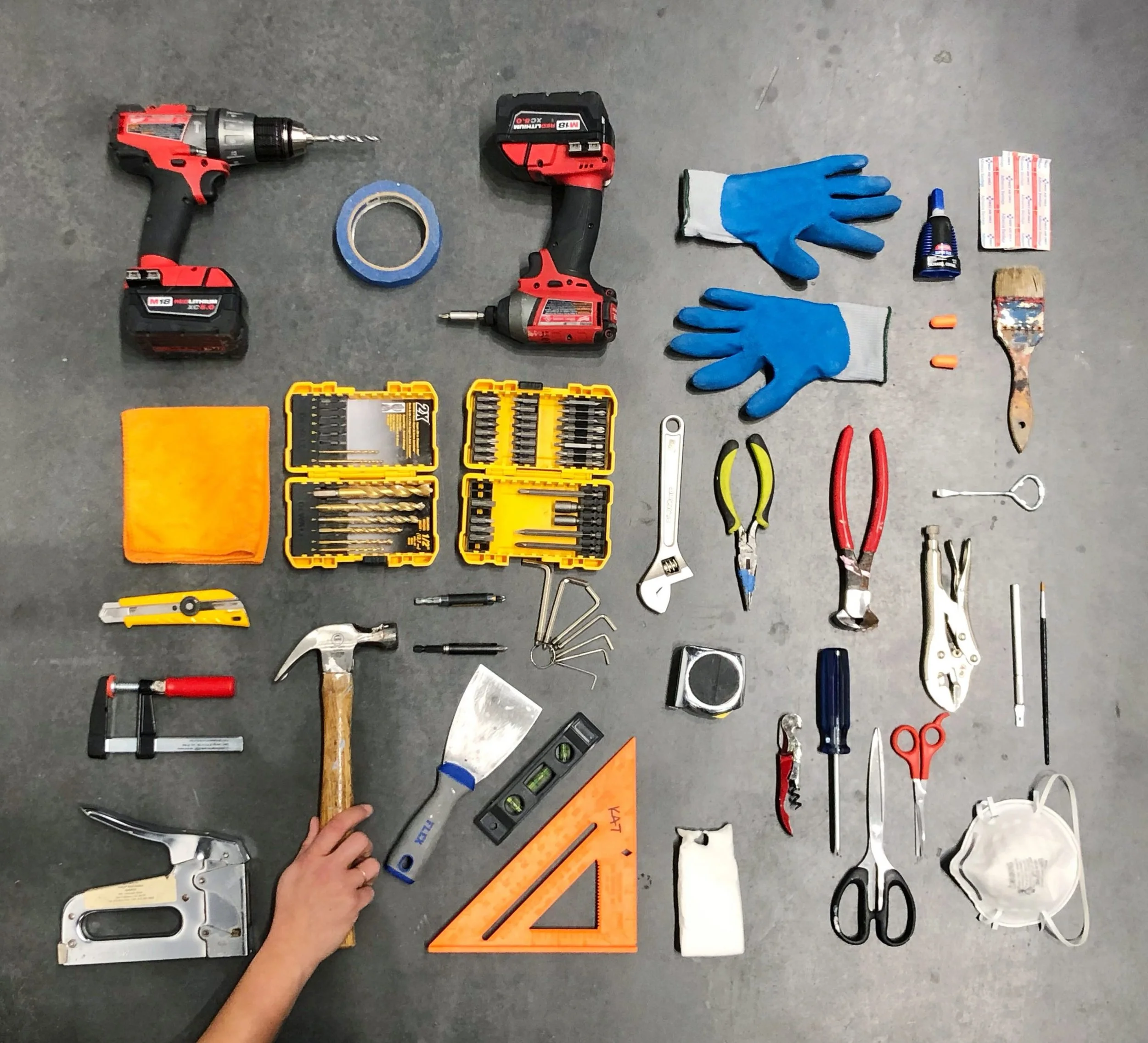 A variety of tools and supplies laid out on a surface, including cordless drills, drill bits, scissors, pens, a level, gloves, painter's tape, a paintbrush, a utility knife, and safety glasses, suggesting a workspace for home improvement or repair projects.