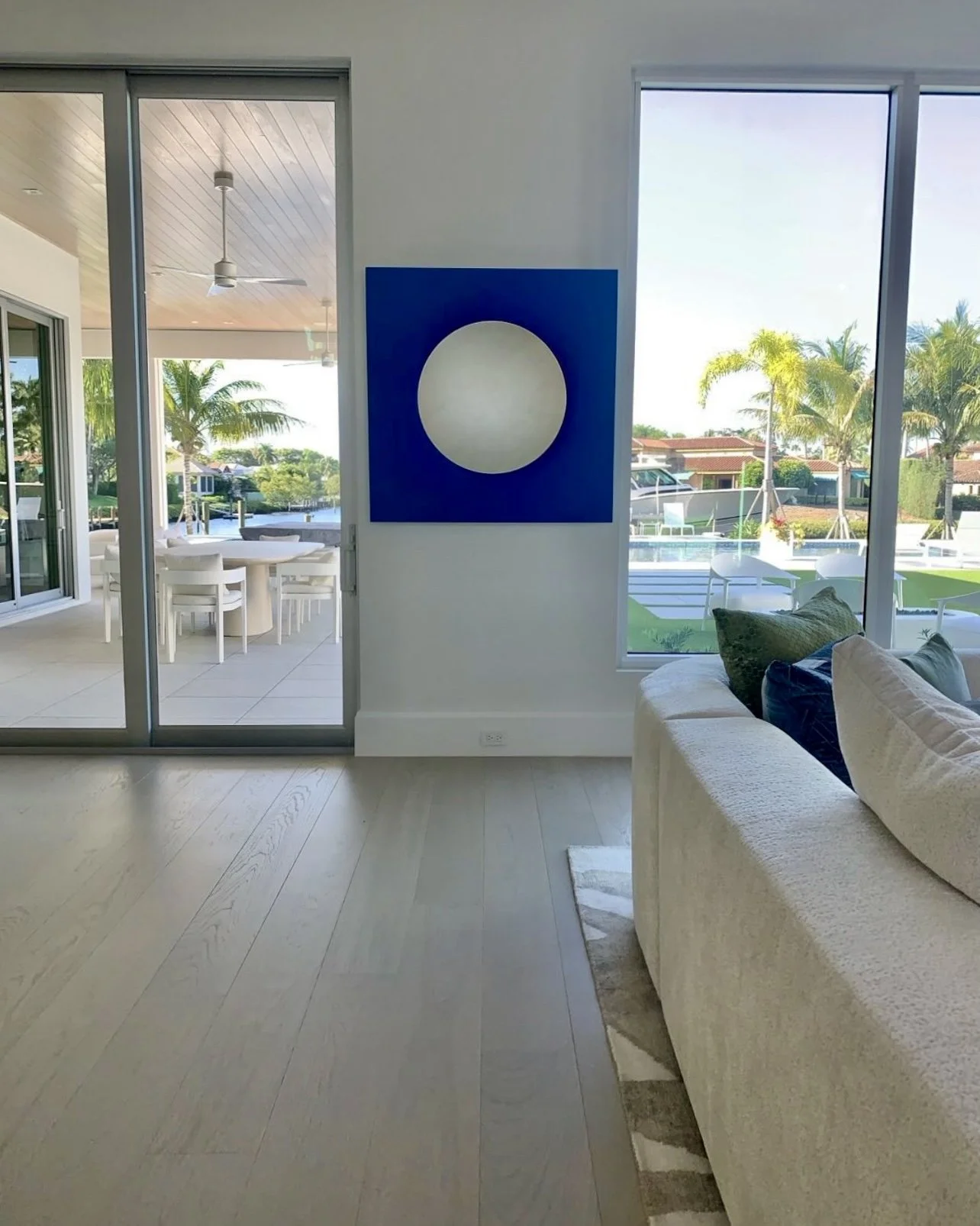 Living room with large window and sliding glass door, view of outdoor patio with dining table and chairs, and landscaped backyard with palm trees.