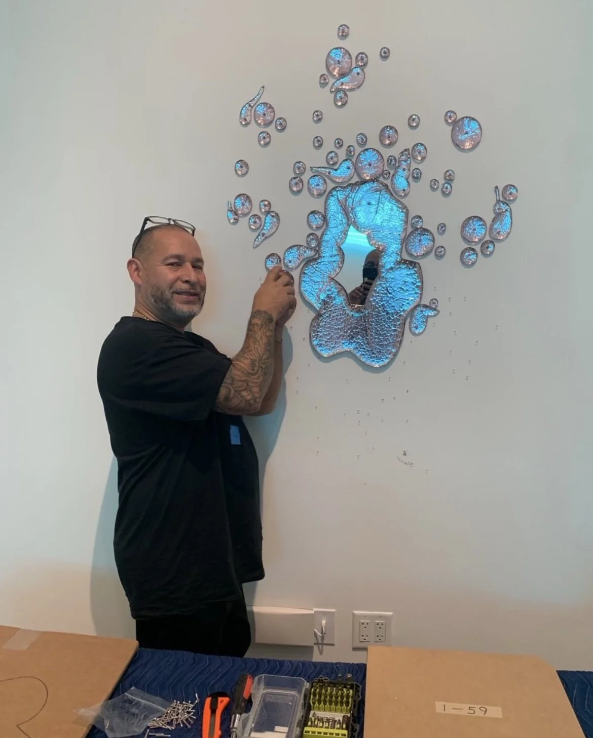 A man with glasses on his head and tattooed arms customizing a decorative mirror on a wall. The mirror is surrounded by droplets of glass or resin with blue lighting, creating a creative art piece.