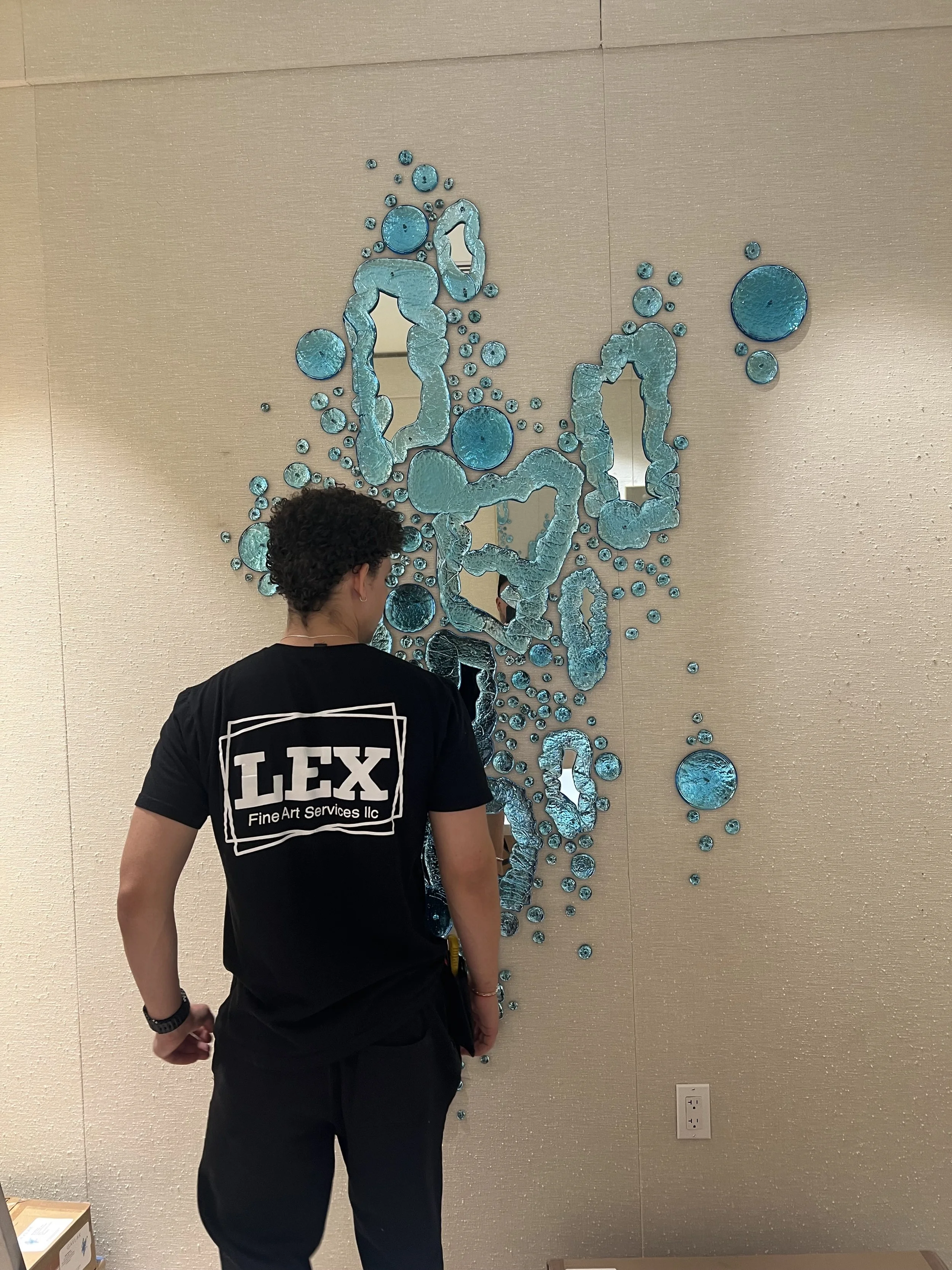 Person with curly hair wearing a black shirt with white text on the back, standing in front of a blue and silver abstract art piece on a beige wall.