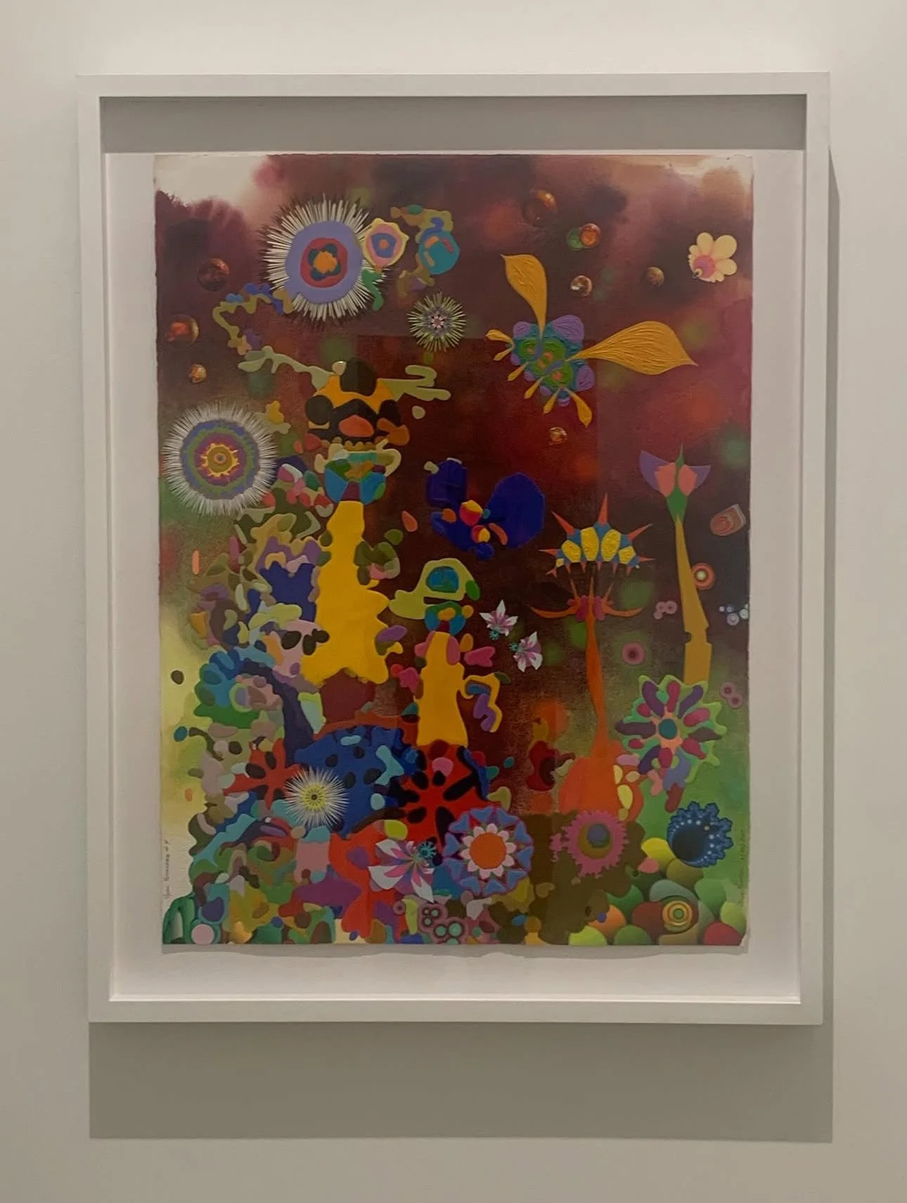 Colorful abstract painting with floral and starburst patterns framed in white, displayed on a white wall.