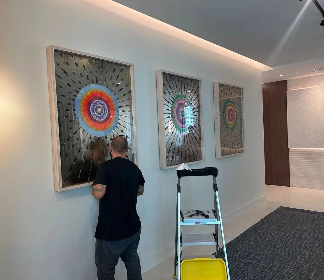 A person with a tattoo on his arm is working on a large framed colorful circular art piece on a white wall. There are three similar art pieces arranged in a row, and a ladder with tools and a yellow container is nearby.