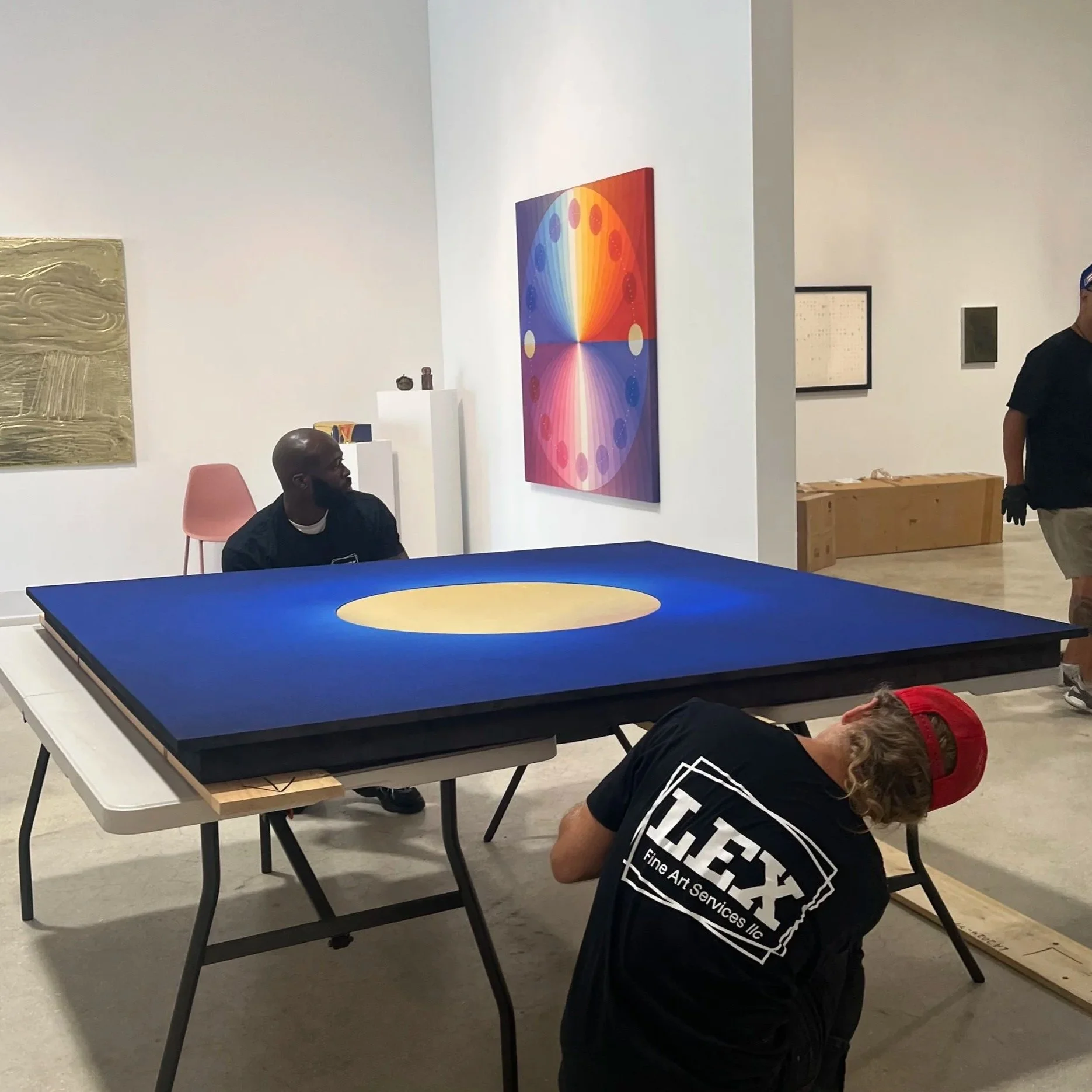 People working on or inspecting a black table tennis table in an art gallery or showroom with white walls, displaying colorful artwork, including a large rainbow-colored geometric painting on the wall.