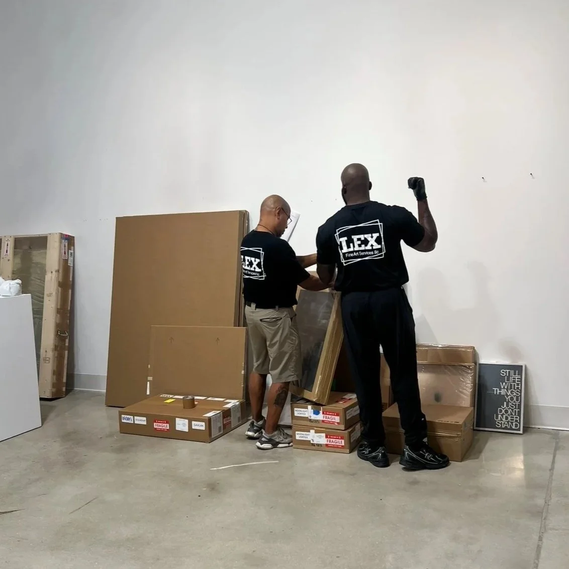 Two men in black shirts moving boxes or furniture in a warehouse or storage space, with cardboard boxes and a framed sign that reads 'STILL LIFE WITH THINGS YOU JUST DONT UNDERSTAND' against a plain white wall.