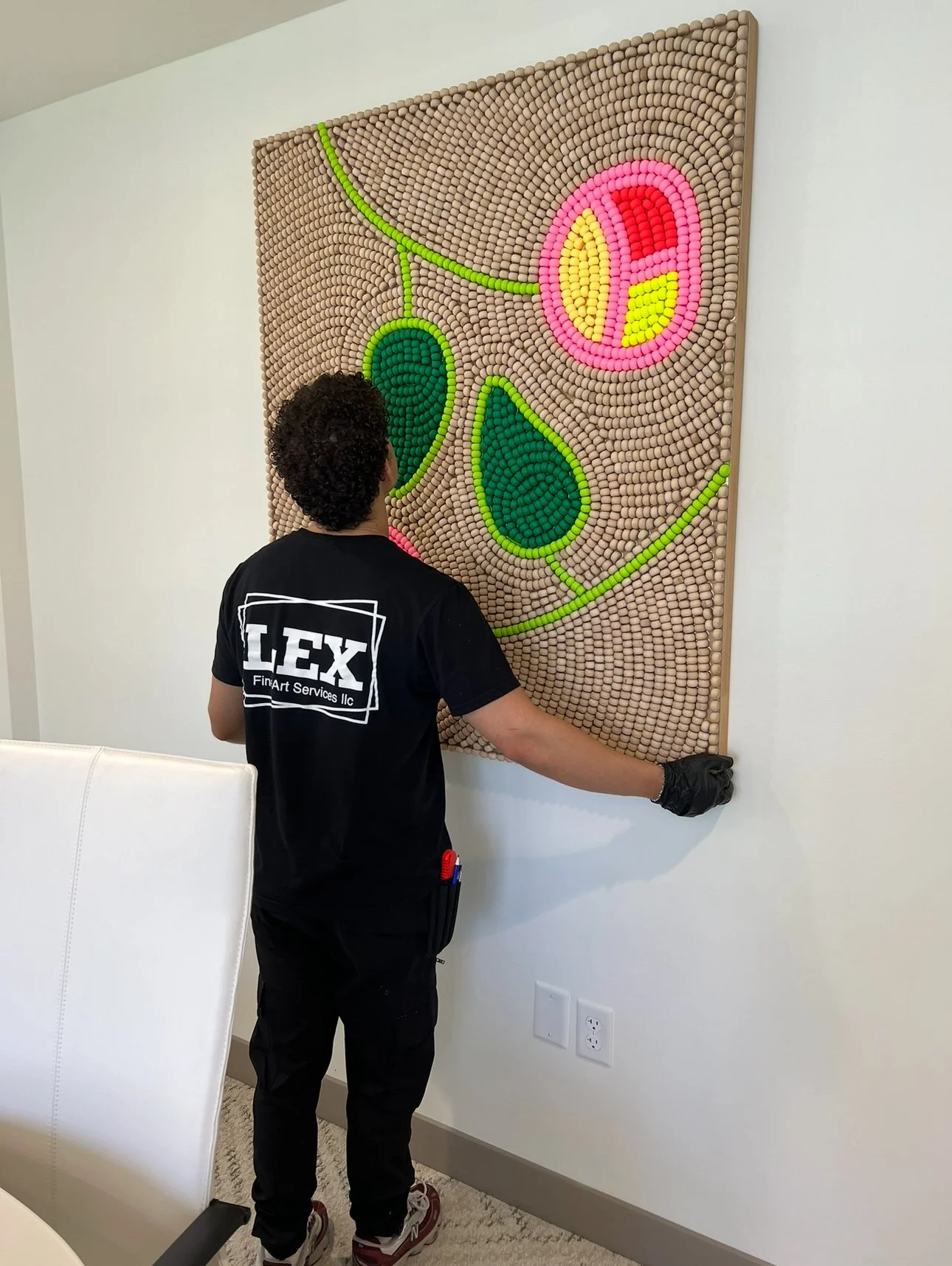 A person wearing black gloves and a black T-shirt with a logo that reads 'LEX FinArt Services LLC' is hanging or installing a large, colorful, textured artwork on a white wall. The artwork appears to be made from numerous small, rounded objects arranged in an abstract floral design with pink, yellow, red, and green colors. The person's face is not visible as they are facing the wall, and they seem to be carefully handling the artwork.