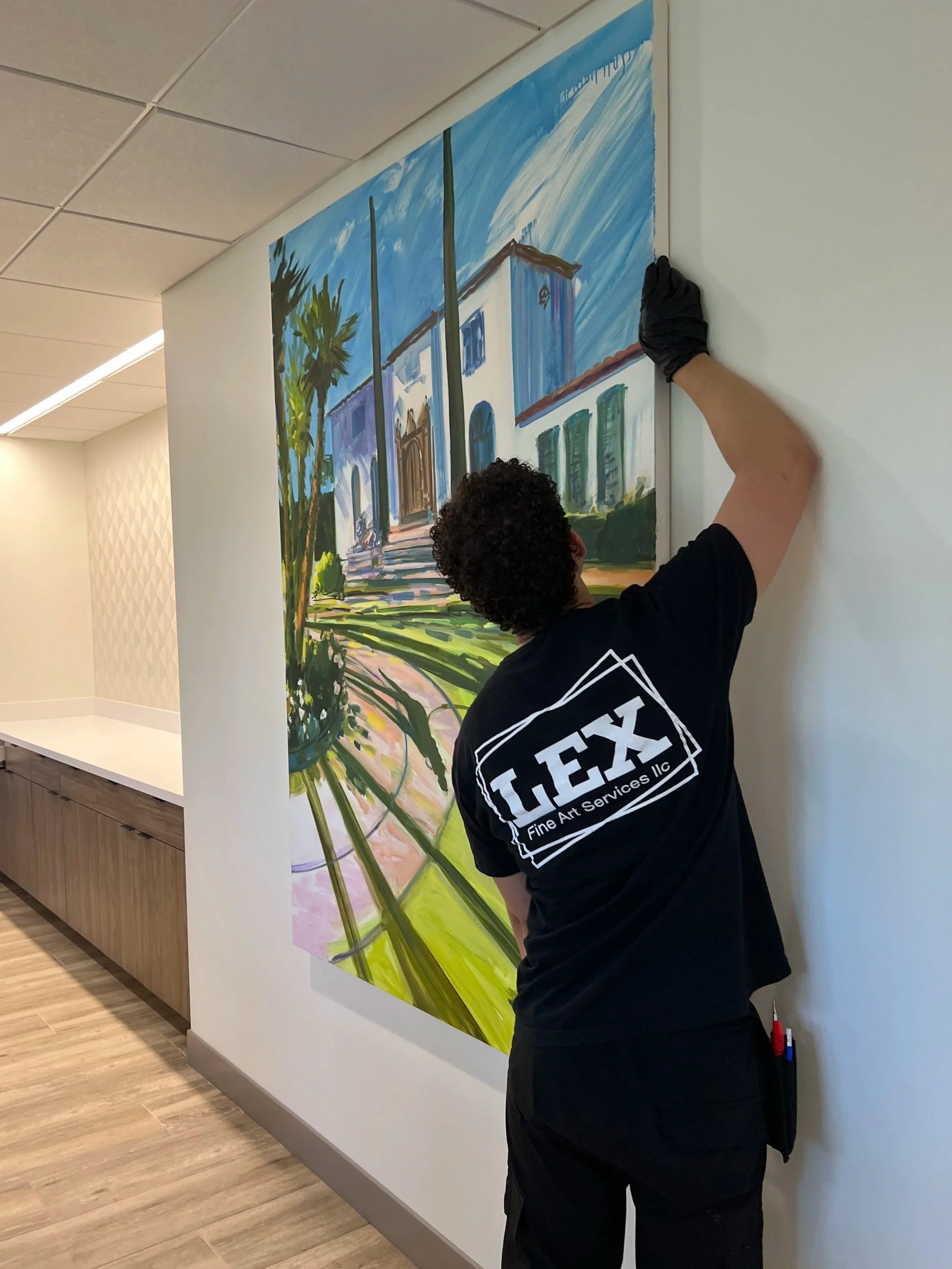 A person with curly hair wearing a black T.E.X. company T-shirt, black gloves, and black pants is hanging a colorful painting of a building with green palm trees on a white wall in an indoor space with beige and wooden flooring.