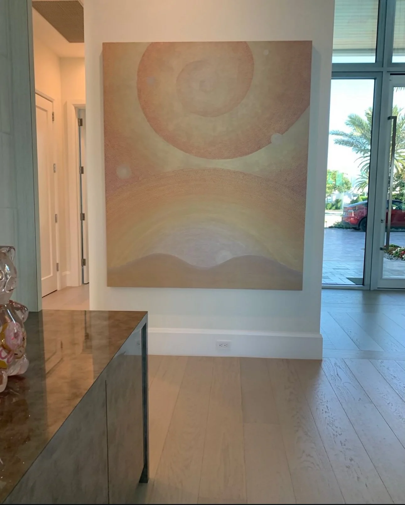 Large abstract painting with swirling pastel colors on a wall inside a modern building with glass doors and windows showing palm trees outside.