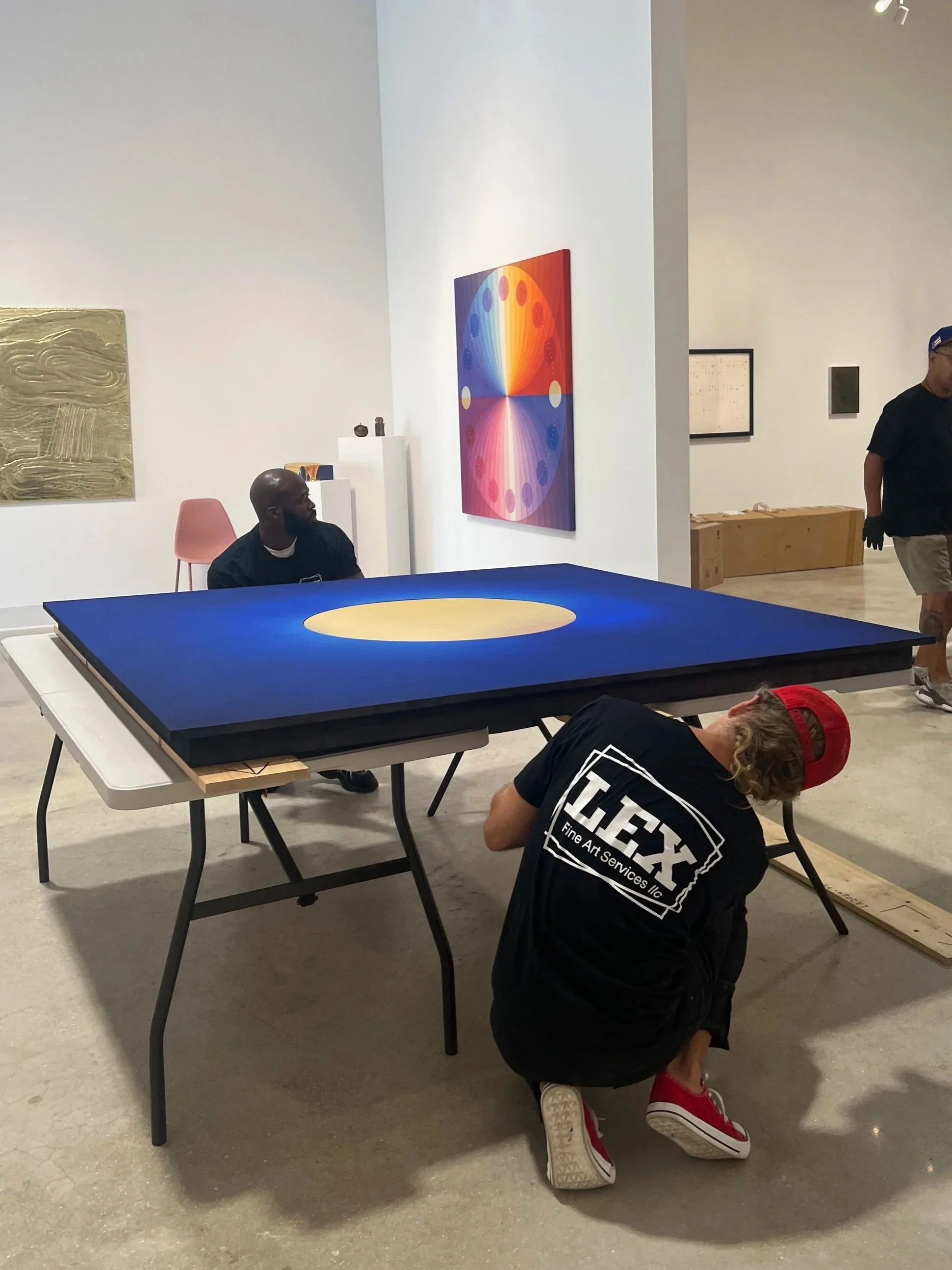 People in an art gallery: one person sitting at a table and two people working on a project, with various artworks hanging on the white walls.