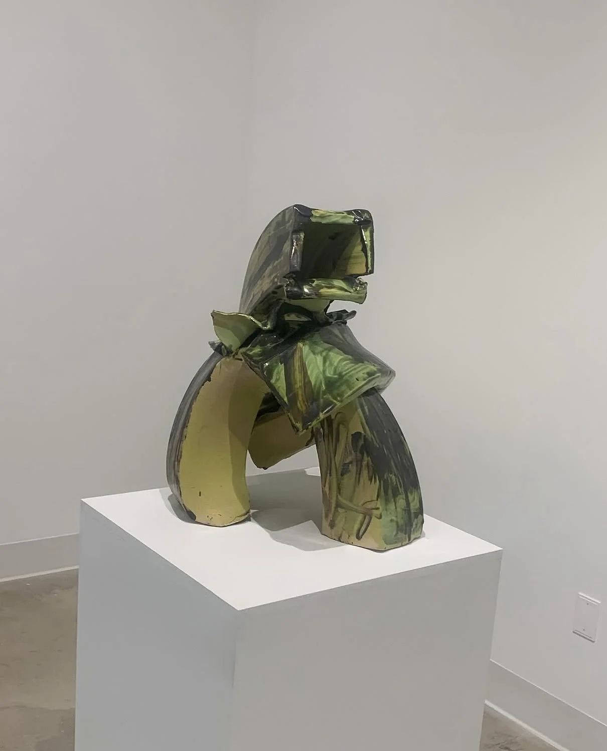 Abstract sculpture made of twisted and layered ceramic or glass pieces with green, black, and yellowish colors, displayed on a white pedestal in an art gallery.