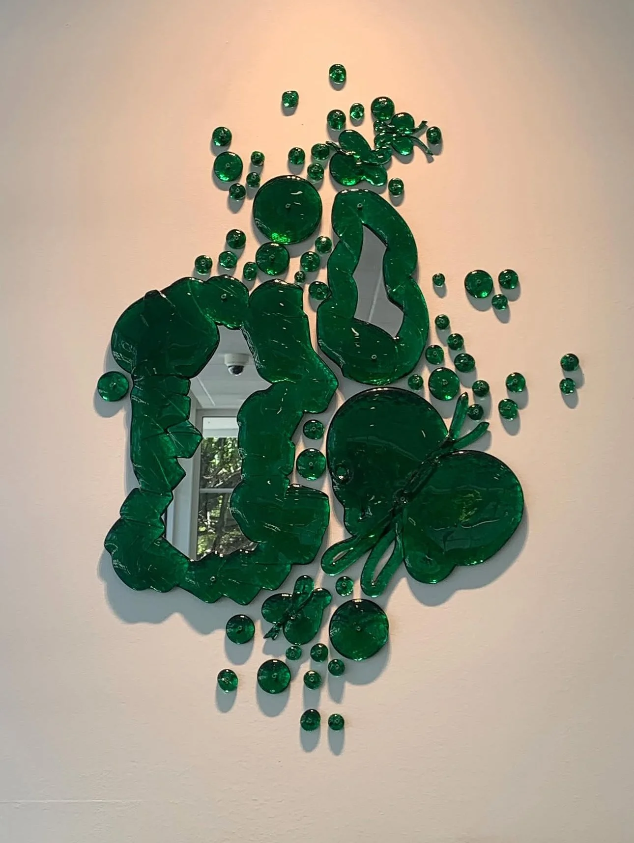 A mirror on a white wall surrounded by green decorative glass pieces shaped like various plants and flowers.