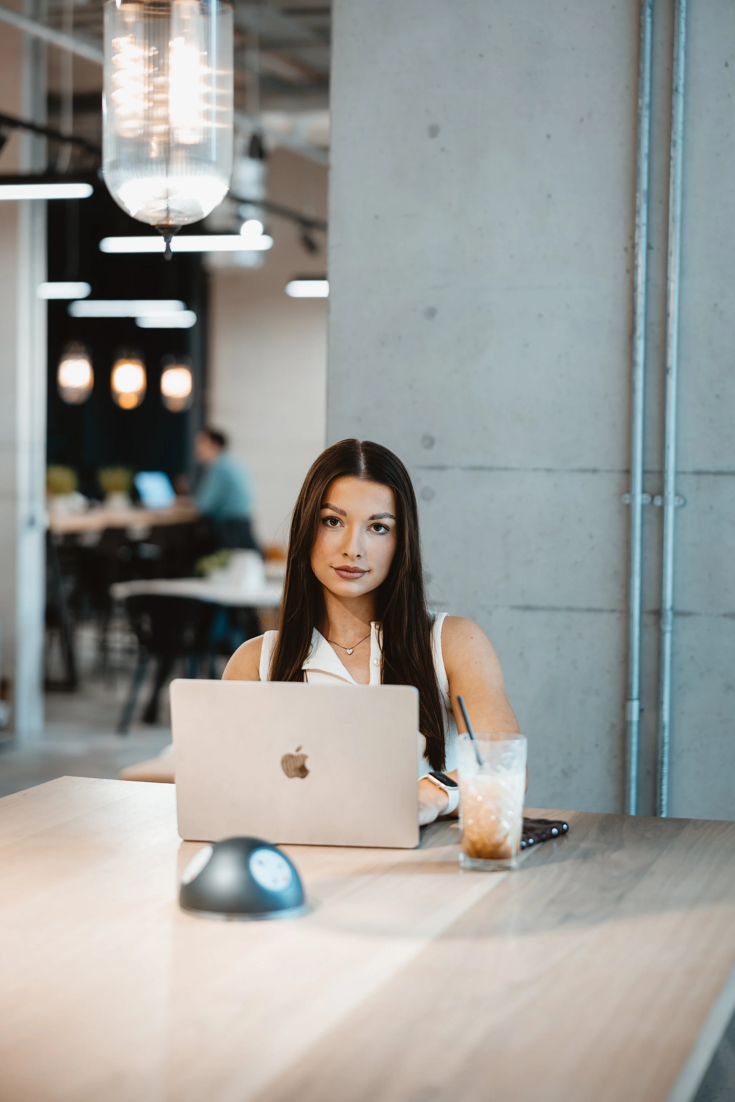 Young woman with long brown hair working on a laptop at a wooden table in a modern office or cafe, with a drink, mouse, and smartphone nearby, and industrial-style decor in the background.
