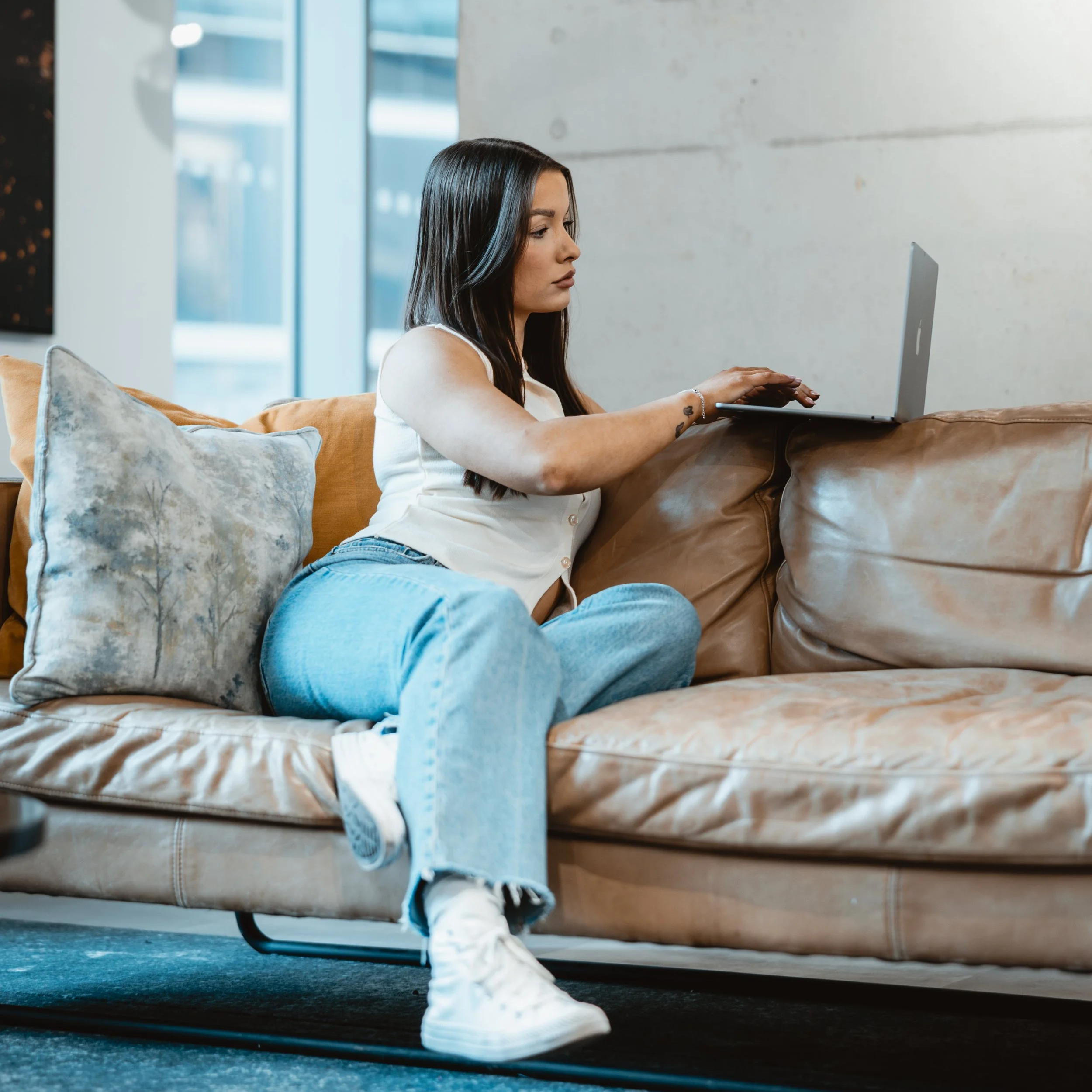 A young woman with long dark hair wearing a white tank top and torn jeans sitting on a beige couch, working on a laptop in a modern, bright room with large windows.