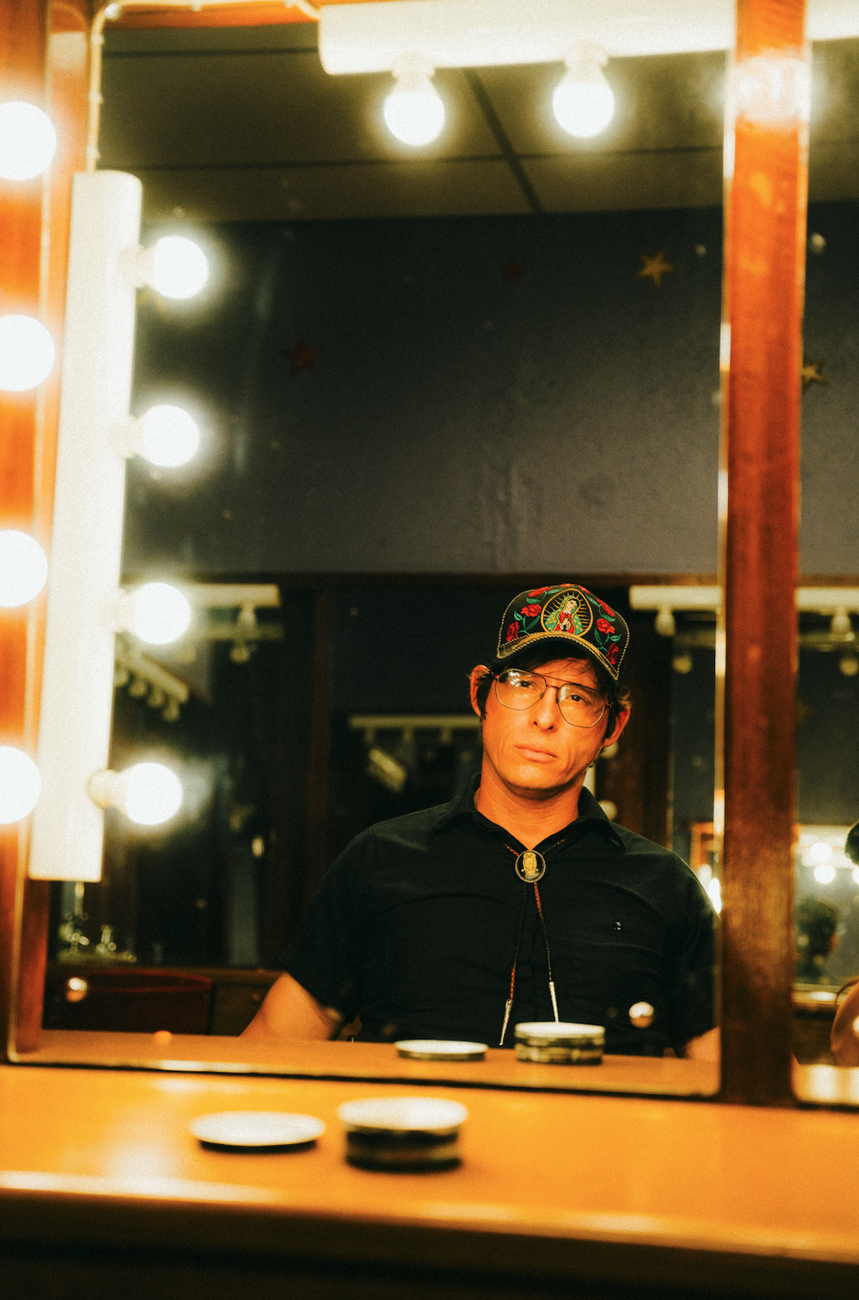Stephen Wilson Junior with glasses wearing a colorful embroidered cap sitting in front of a mirror with bright light bulbs, with a reflective and contemplative expression.