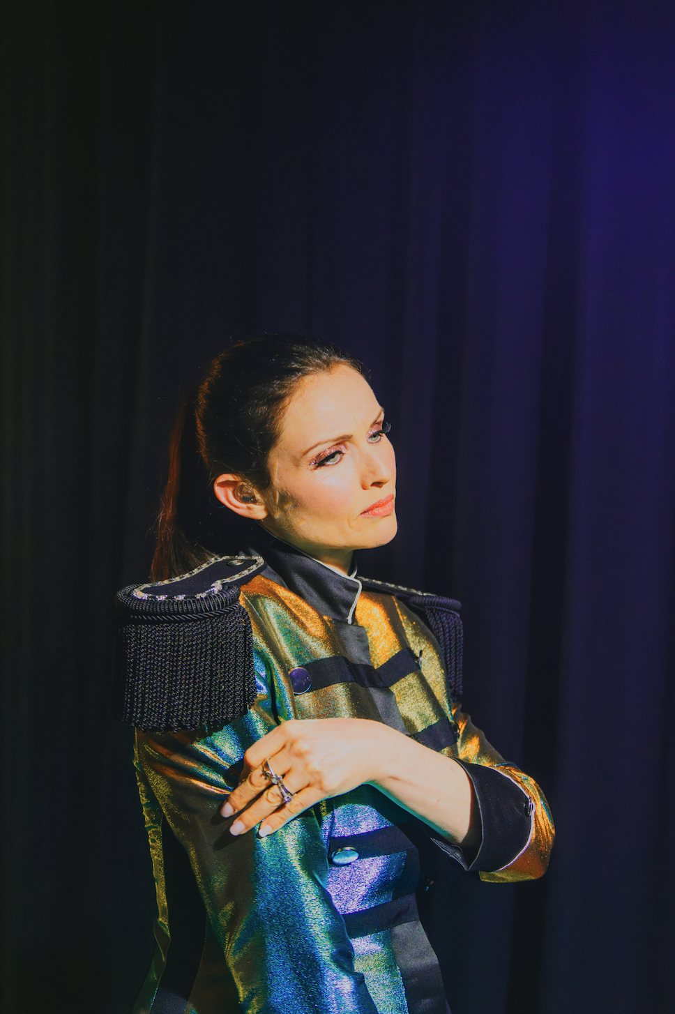 Sophie Ellis Bextor dressed in a colorful, shiny jacket with black lapel and shoulder epaulets, standing against a dark purple background, looking to the side with a serious expression.
