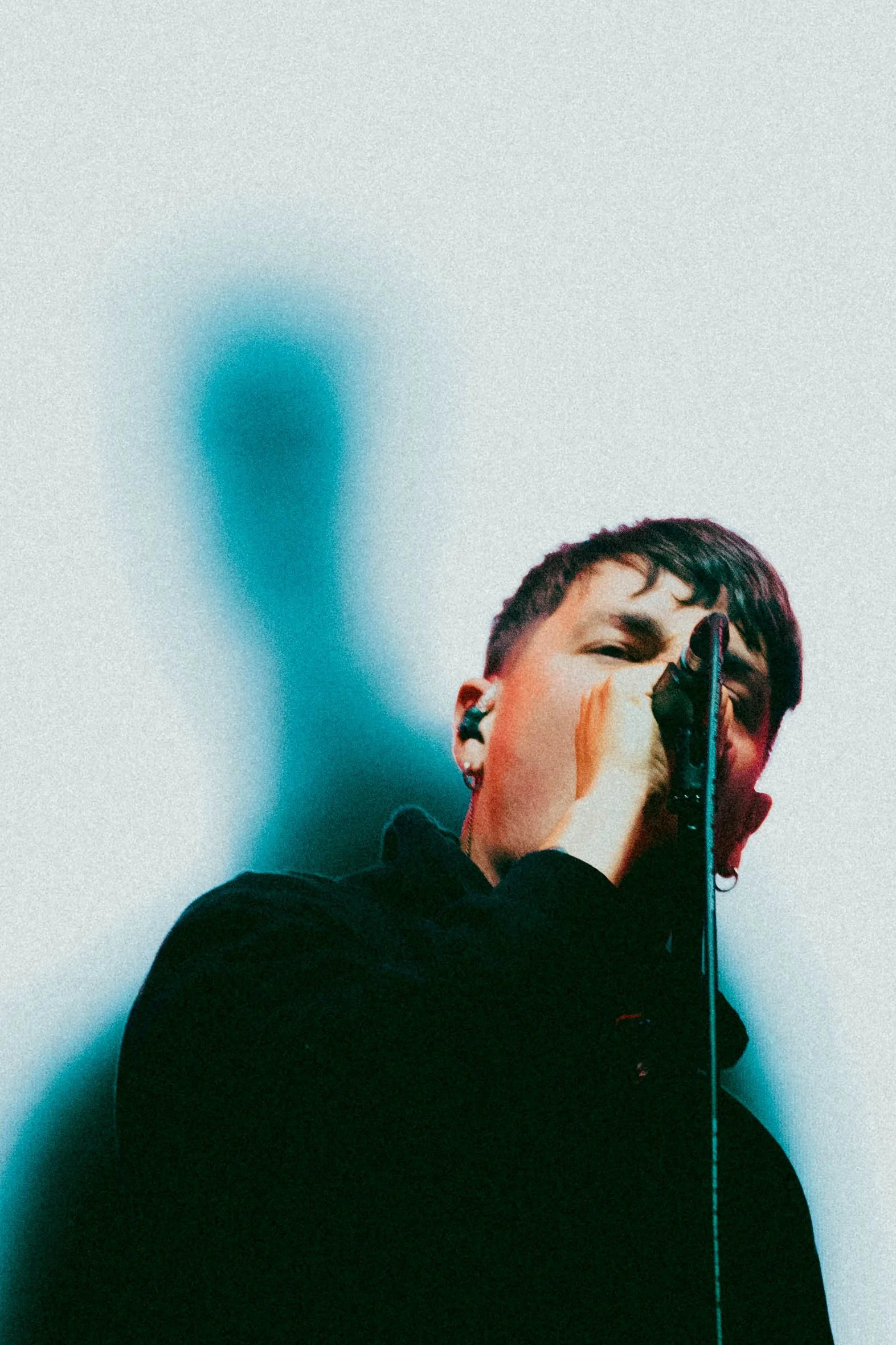 Boston Manor Singer A person with short dark hair holding a microphone close to their mouth, wearing a black top, against a plain light-colored background.